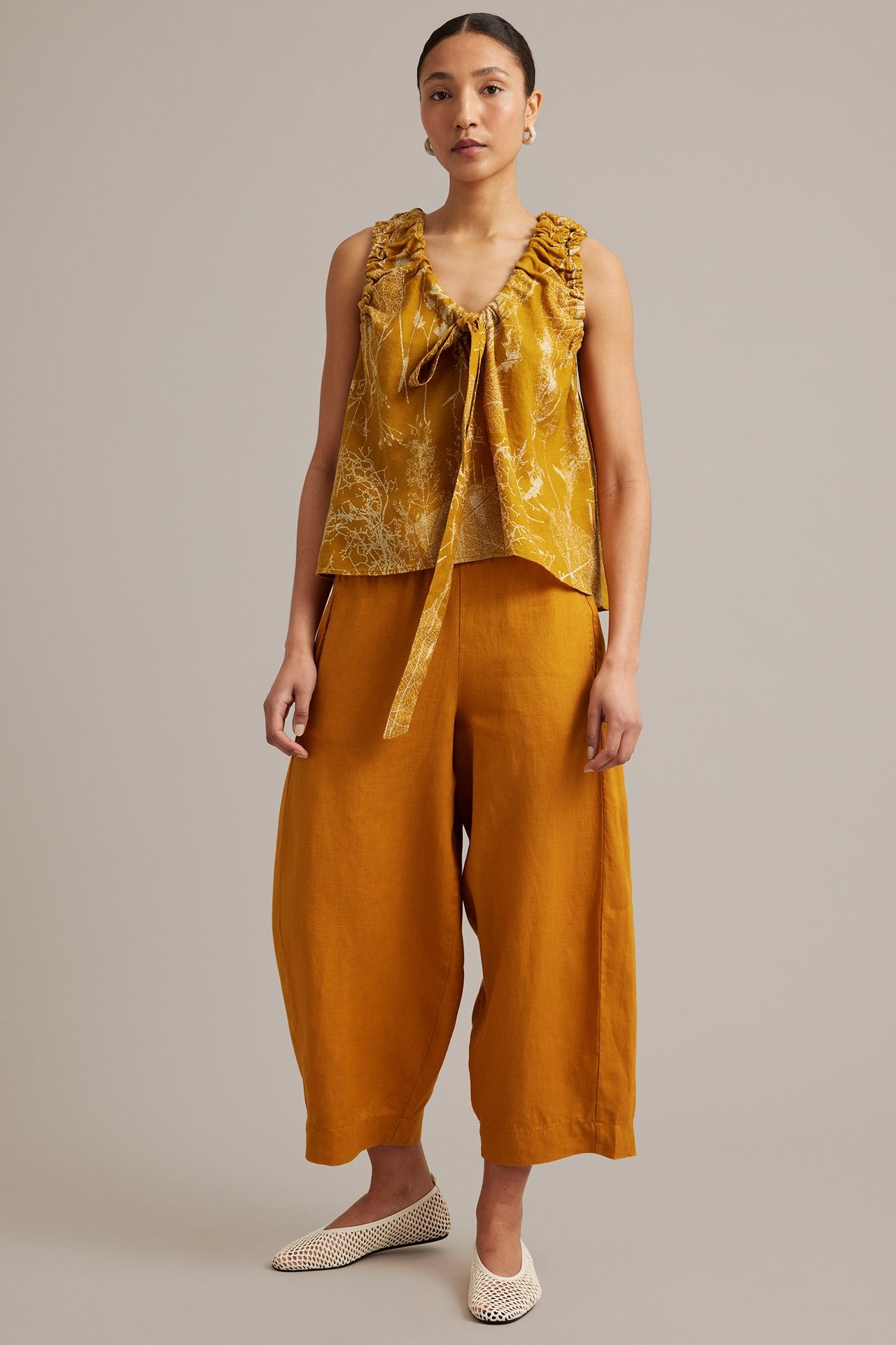 Framifrån, kvinna i lös gulblommig linnetopp med knytband, matchande vida linnebyxor och cremefärgade nätballerinor.