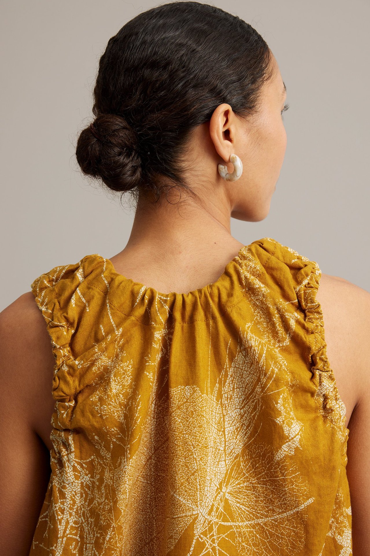 Bakifrån: kvinna i senapsgul linnetopp med vitt bladmönster och ljusa marmorerade ringörhängen.
