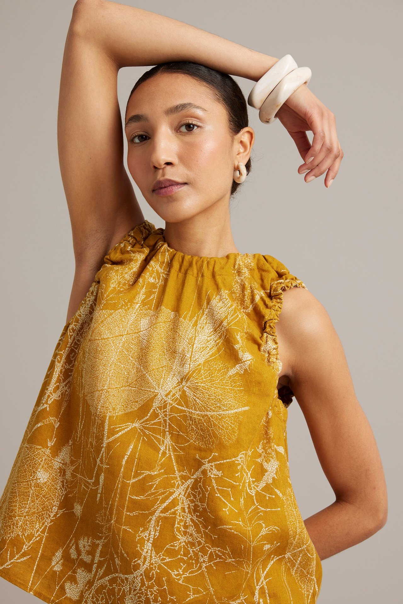 Närbild framifrån på kvinna i gul linnetopp med vitt bladtryck, ljusa ringörhängen och armringar.