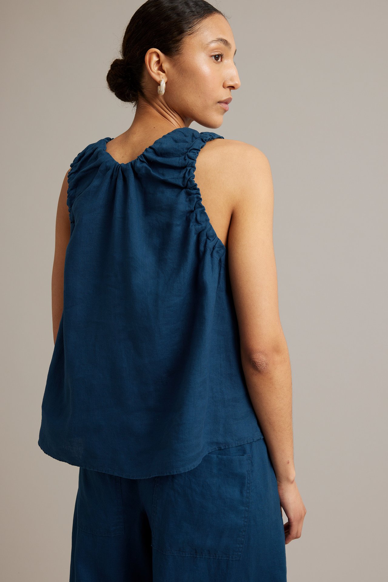 Sidanvy av kvinna i lös blå linnetopp med rynkad ringning, matchande blå byxor och ringörhängen.