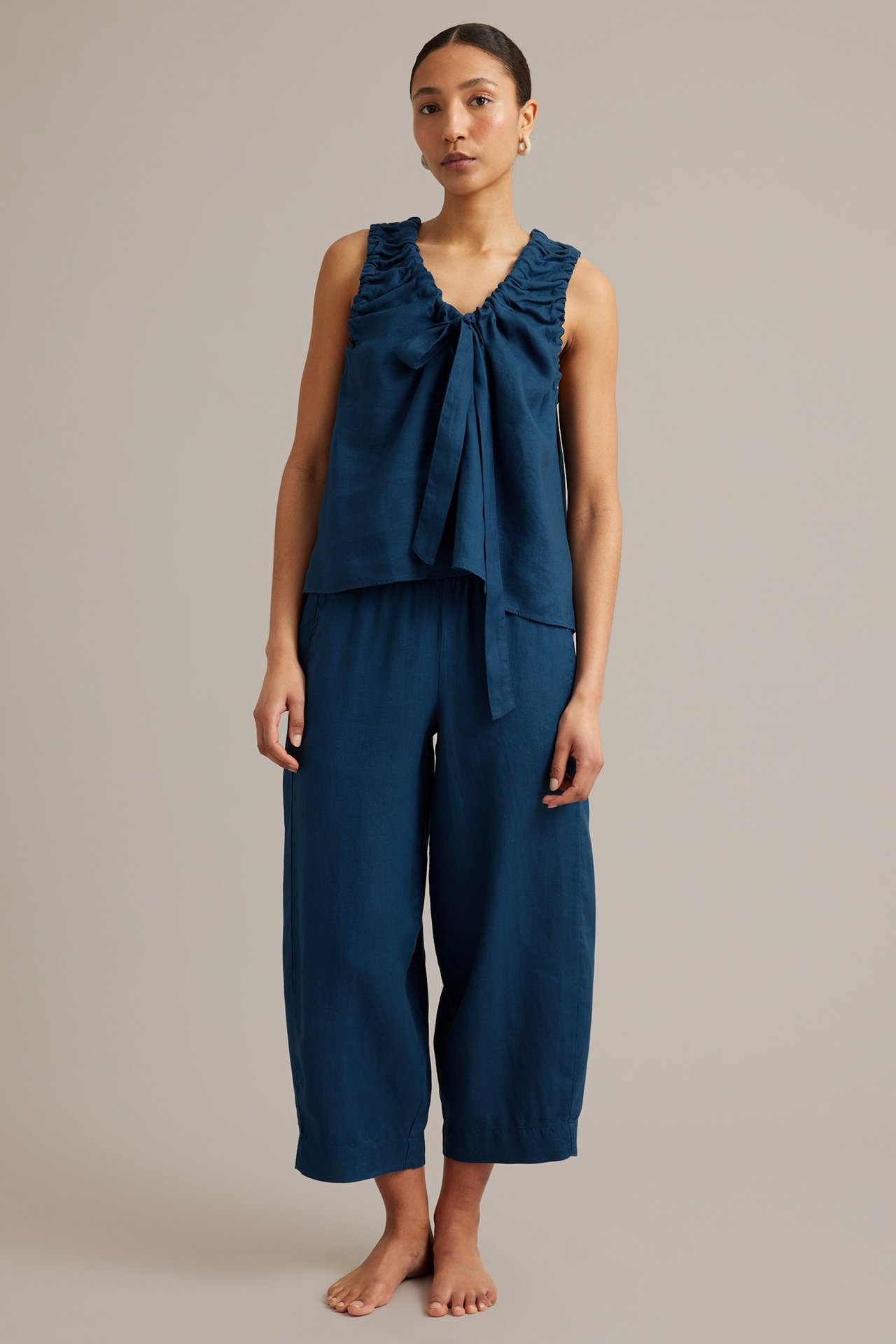 Framifrån, kvinna i löst sittande blå linnetopp med knytning i ringning och matchande vida, korta byxor.
