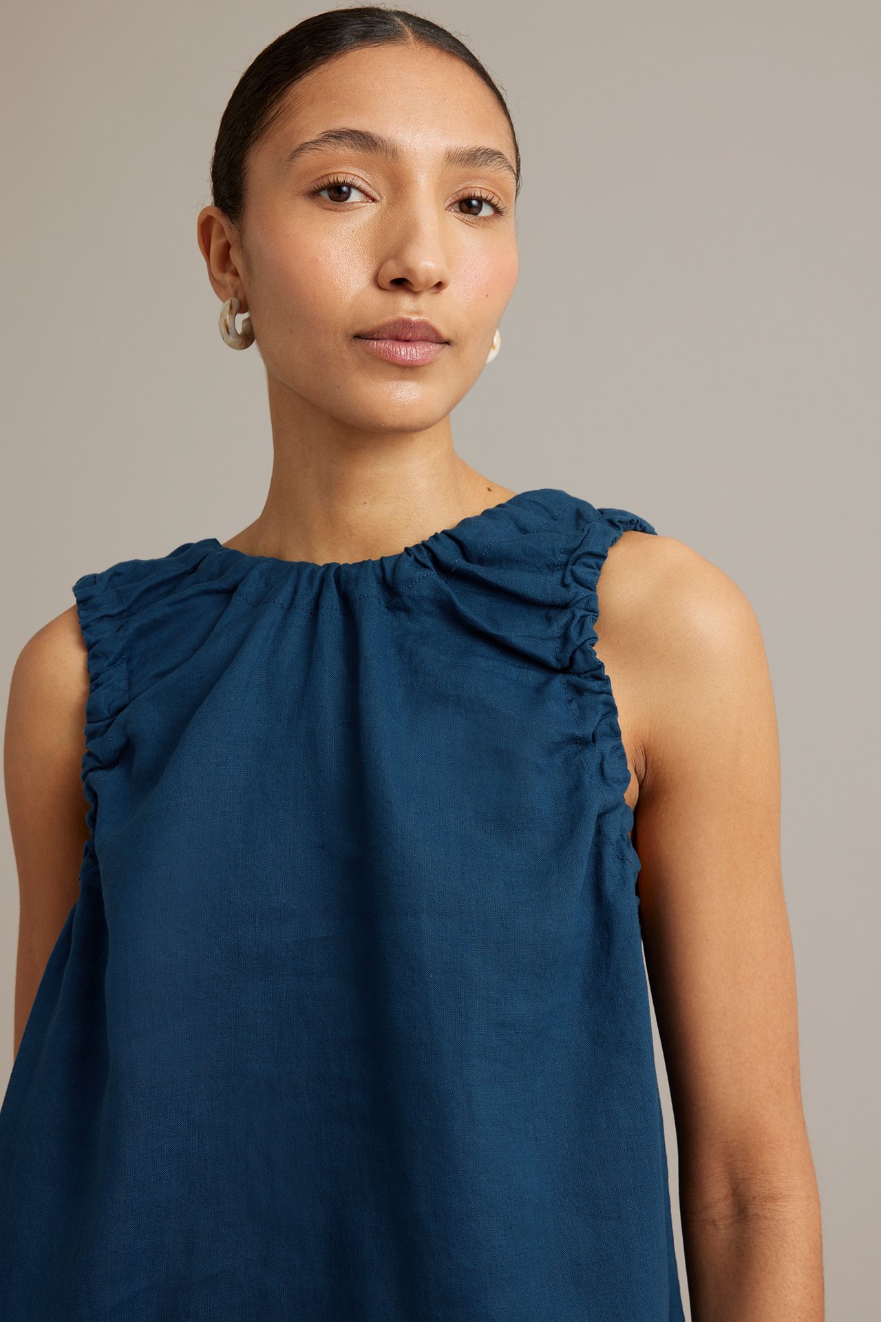 Närbild framifrån, kvinna i blå linnetopp med rynkad ringning och axlar, bär krämfärgade ringörhängen.