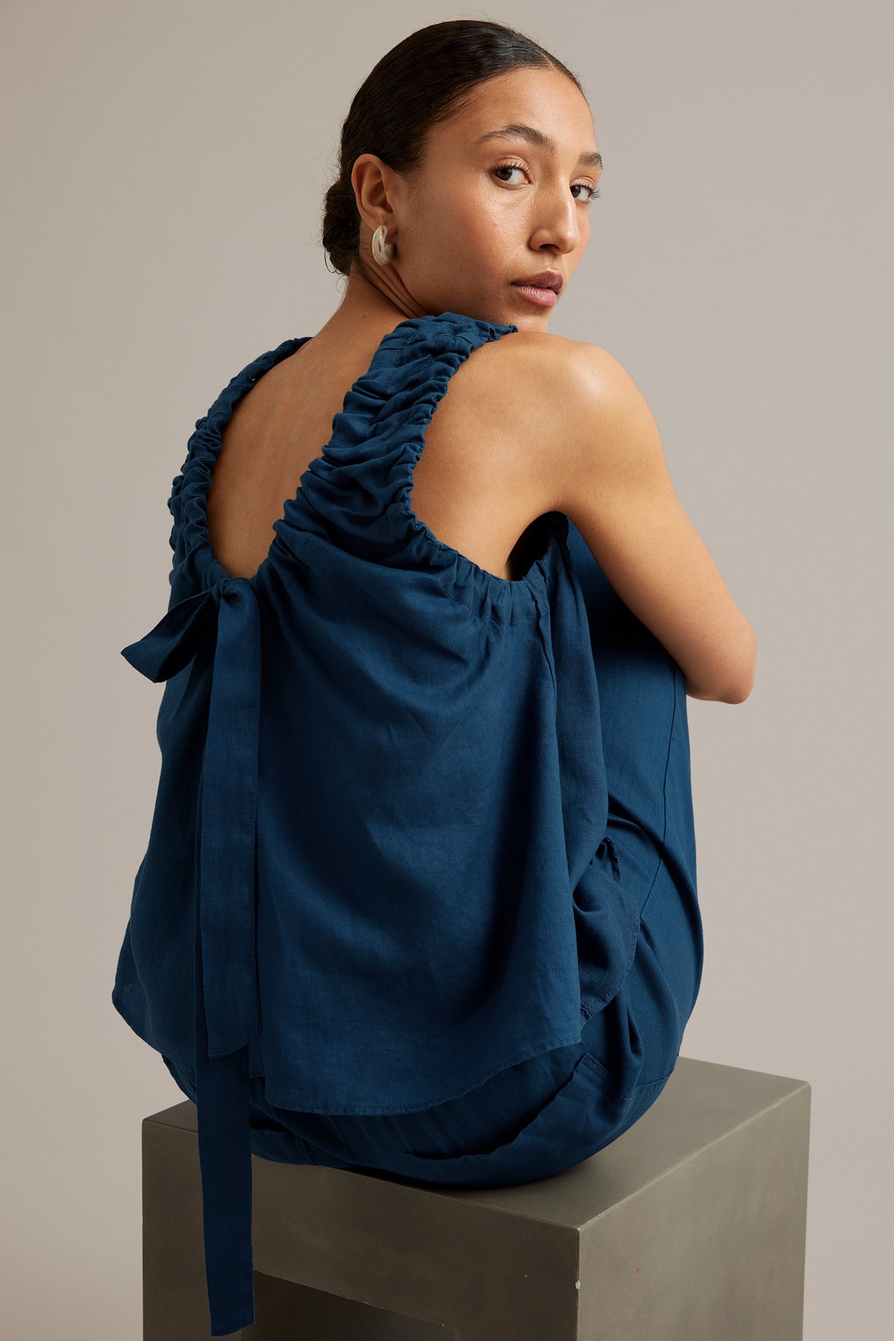 Bakifrån, en kvinna i blå linnetopp med rynkade band och knytning i ryggen, stylad med blå byxor.