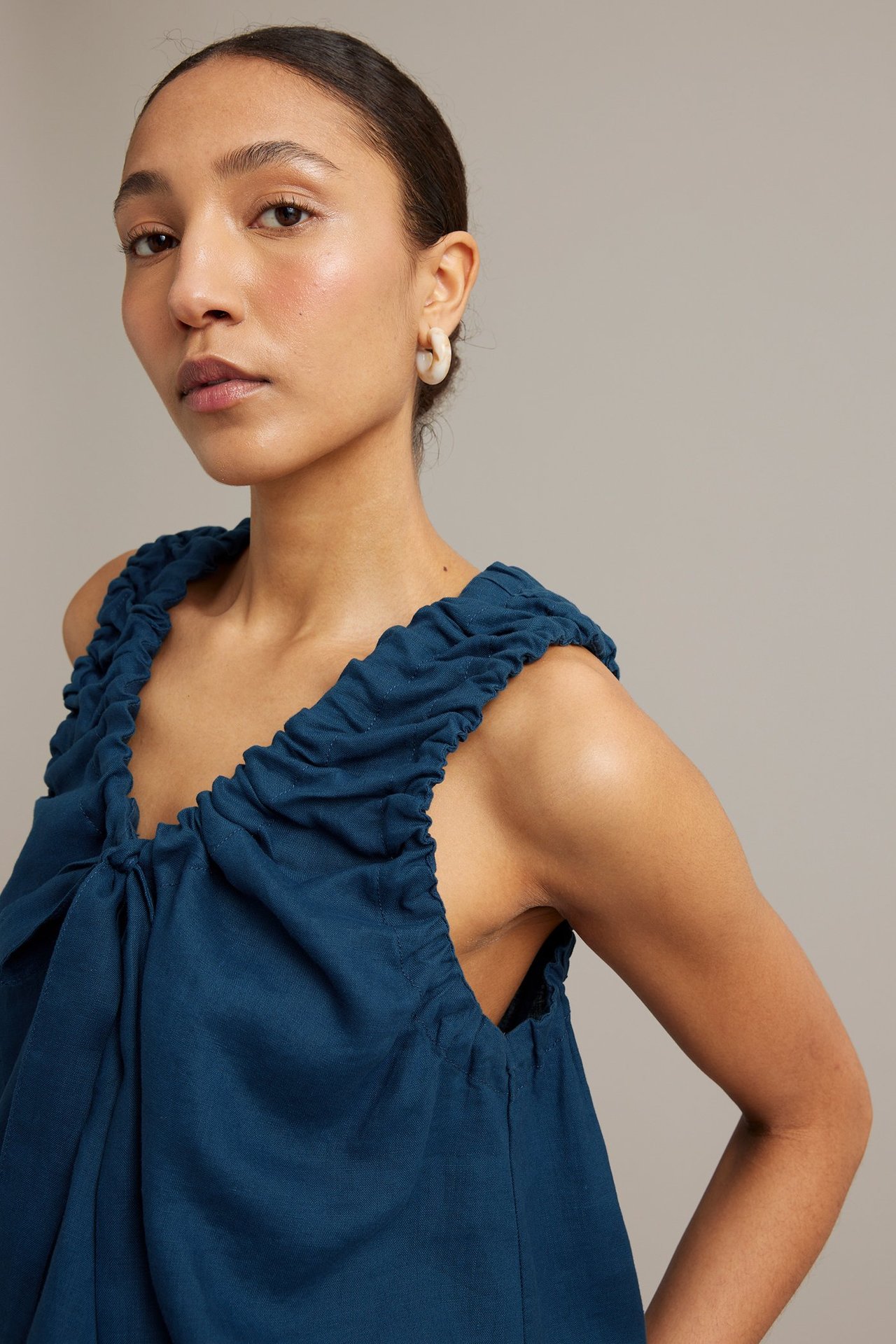 Närbild av kvinna i blå linnetopp med rynkad ringning och knytband, med vita ringörhängen.