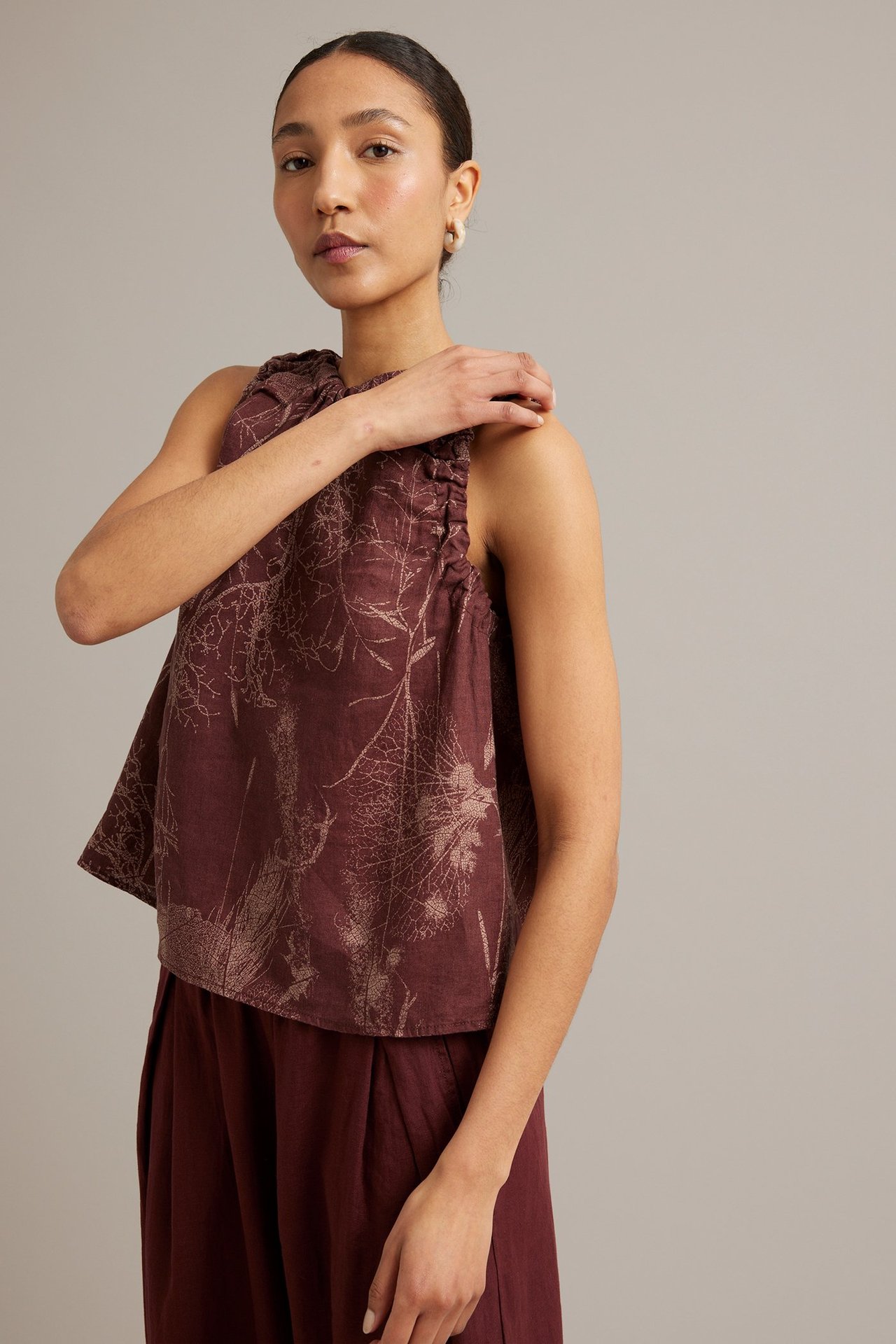 Kobieta w bordowej lnianej kwiecistej bluzce z dekoltem wiązanym, do tego pasujące spodnie. Widok z przodu.