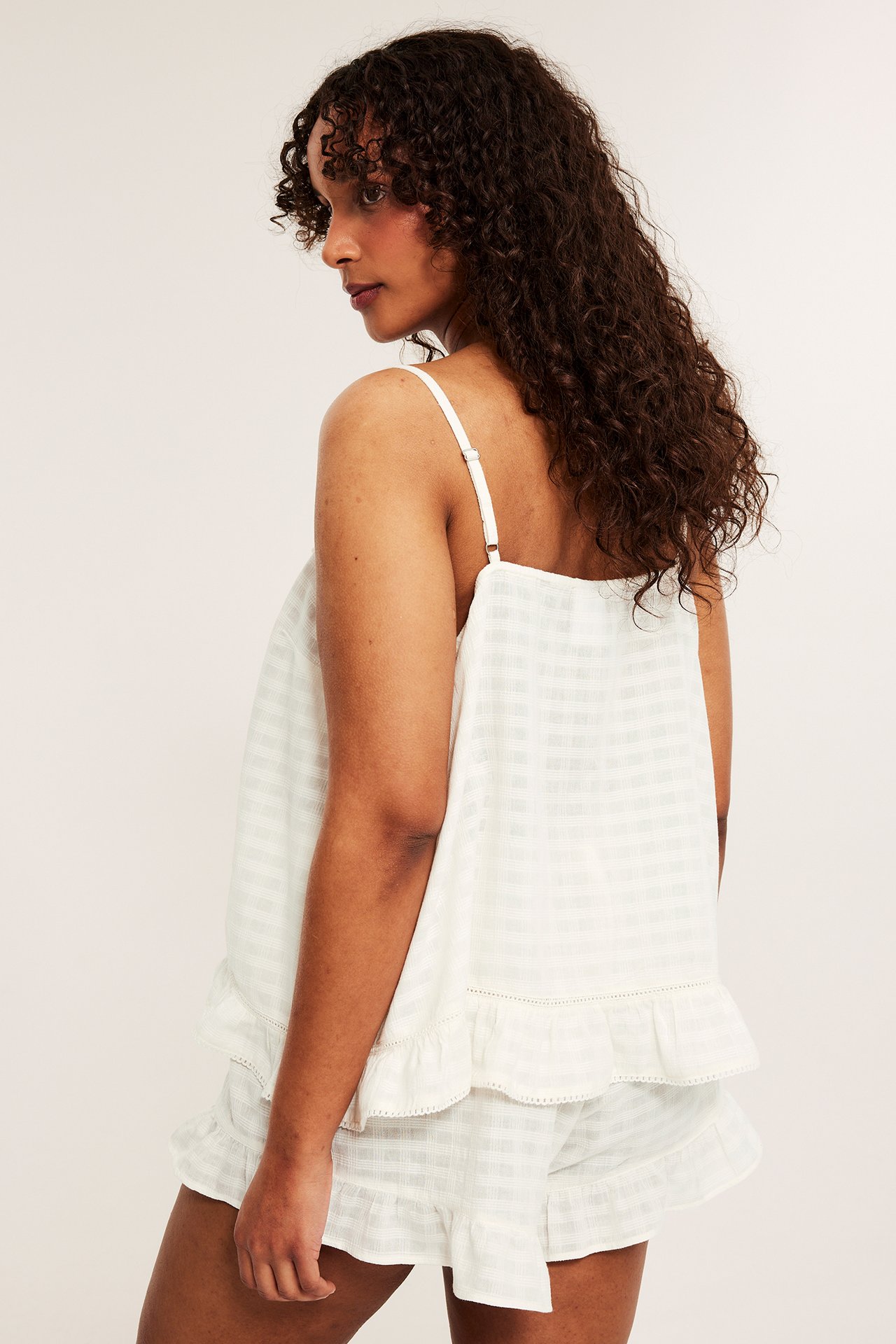 Sidebilde av kvinne i offwhite pyjamas-camicoletopp og matchende shorts i strukturert bomull med rysjekant.