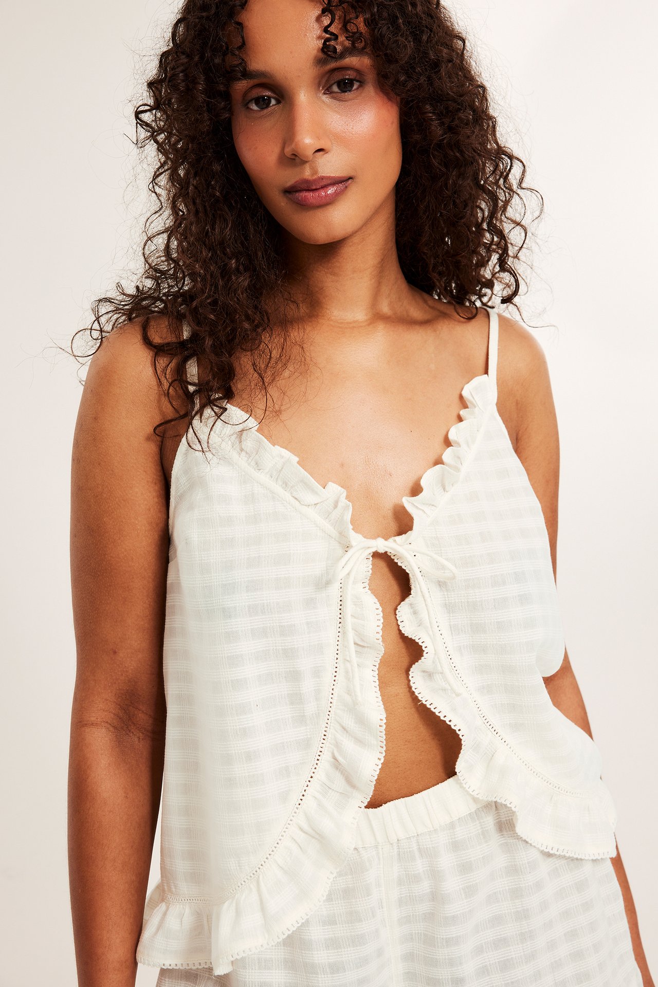 Forfra: Kvinne i offwhite strukturert bomulls-pyjamastopp med knyting, volanger og matchende shorts.