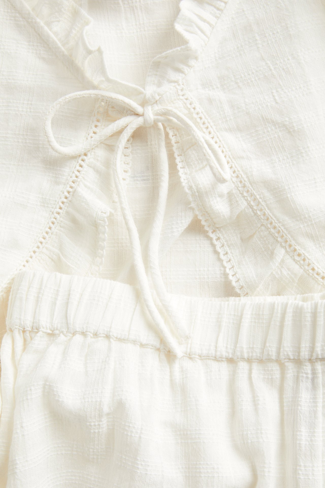 Nærbilde av offwhite pyjamatopp i strukturert bomull med knytebånd foran og rysjekanter, over matchende shorts.