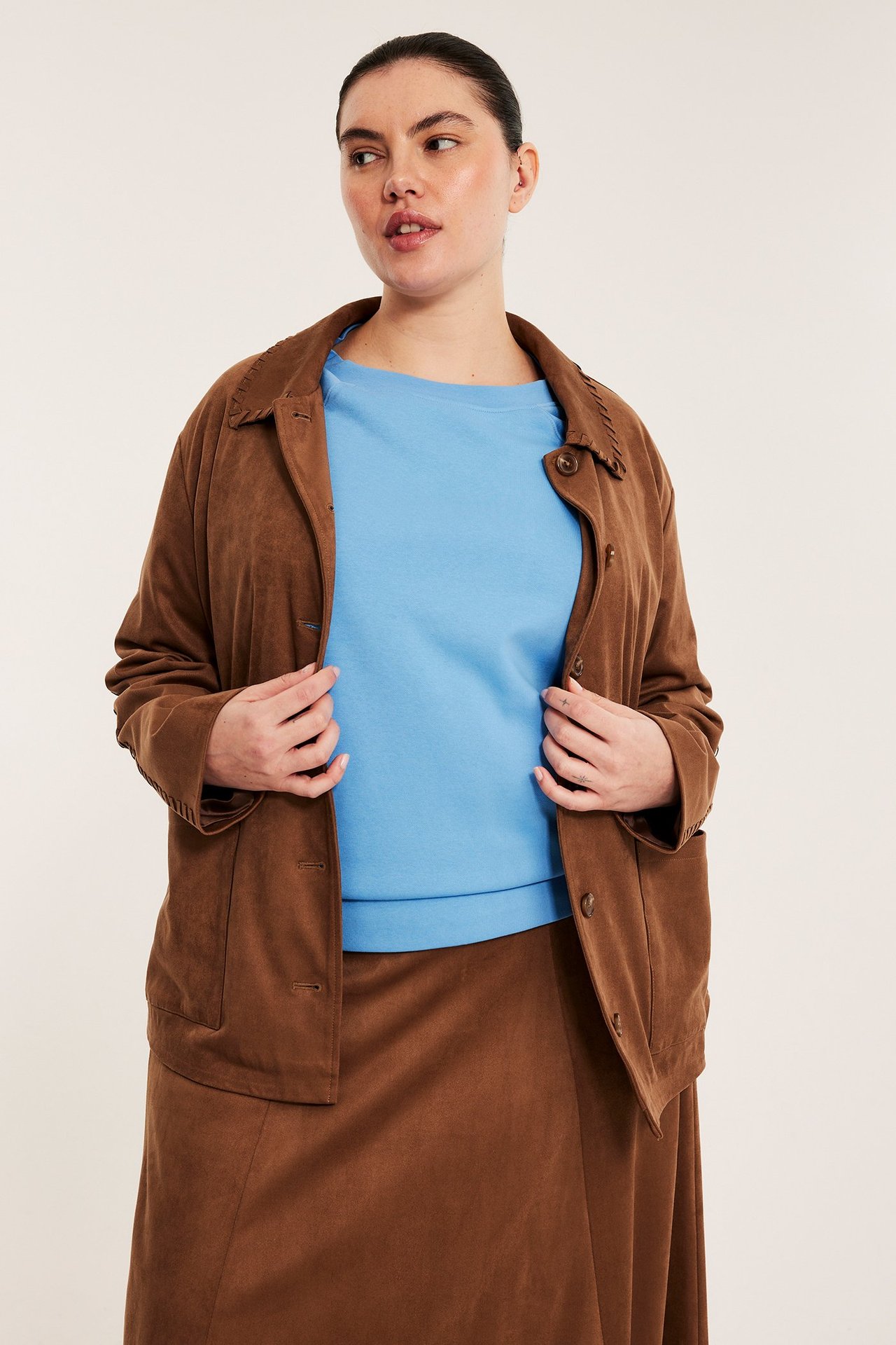 Przód: Kobieta w jasnoniebieskiej bluzie z krótkim rękawem, otwartej brązowej kurtce i brązowej spódnicy, trzyma klapy.