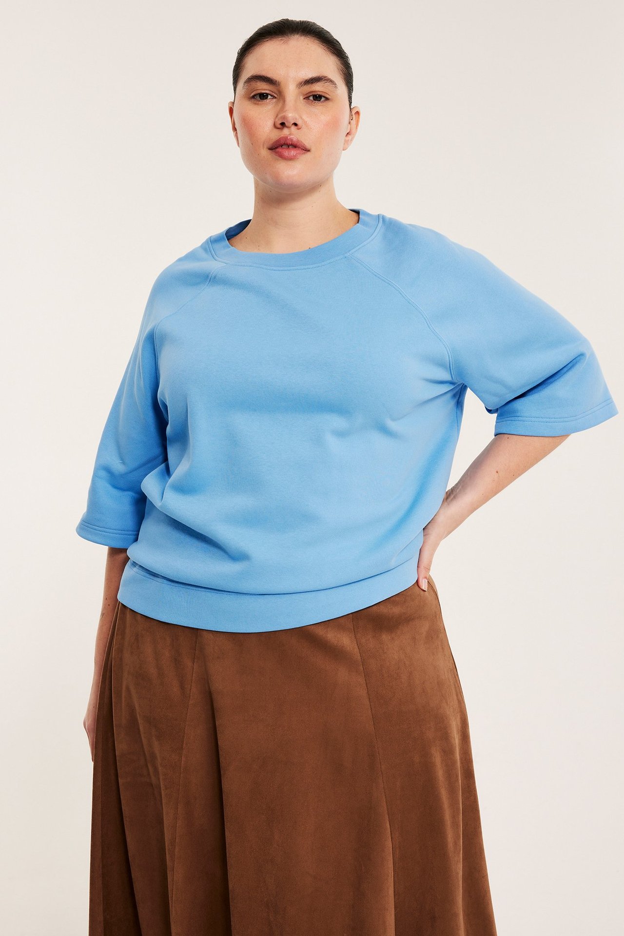 Kobieta przodem w jasnoniebieskiej bluzie z krótkim rękawem i brązowej spódnicy trapezowej.