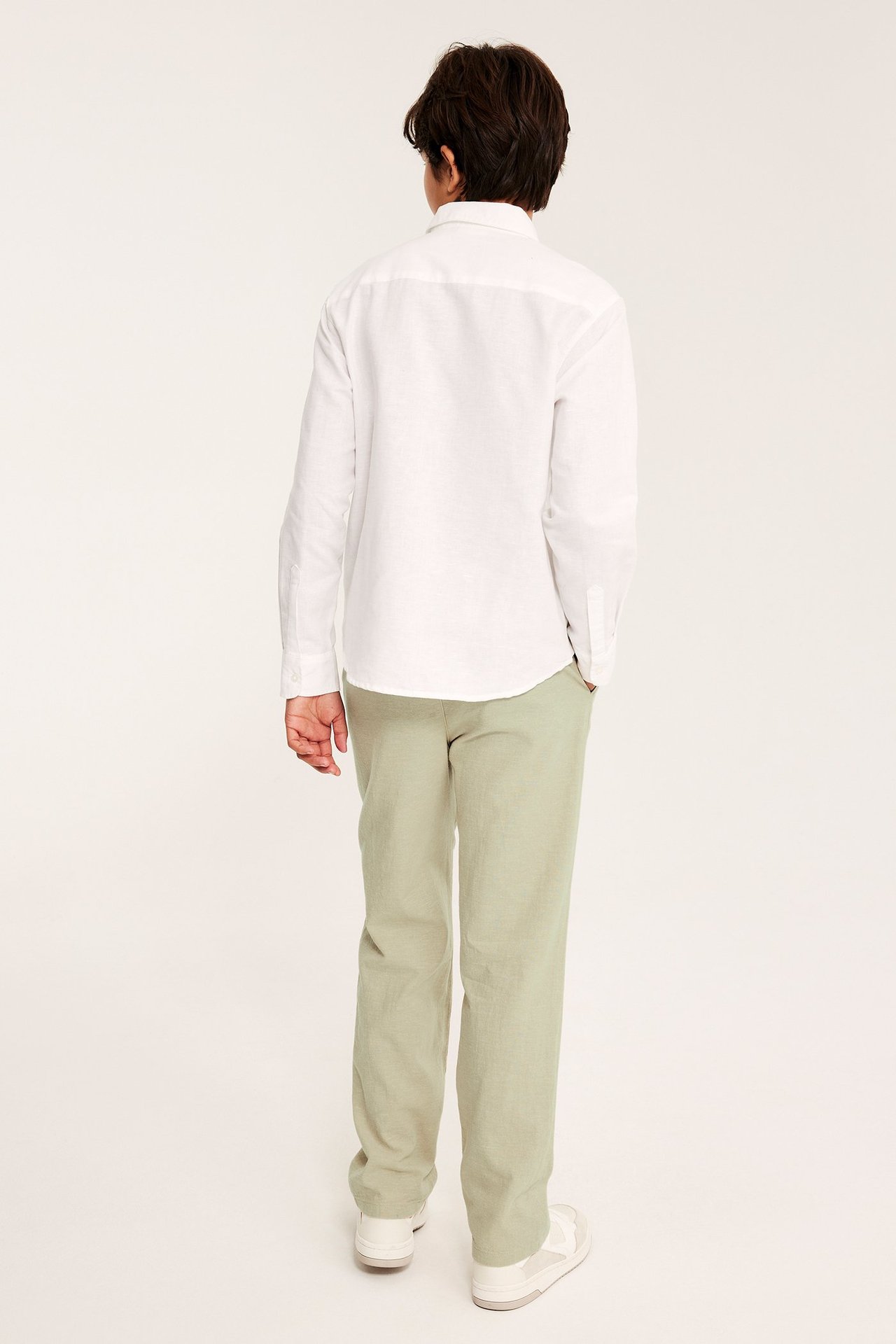 Dziecko, tyłem, w jasnozielonych, luźnych spodniach lnianych, białej koszuli i białych trampkach.