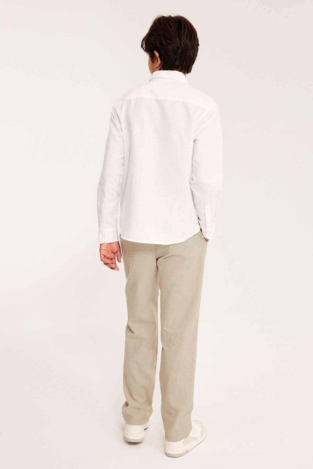Widok od tyłu dziecka w luźnych beżowych spodniach lnianych i białej koszuli z długim rękawem.