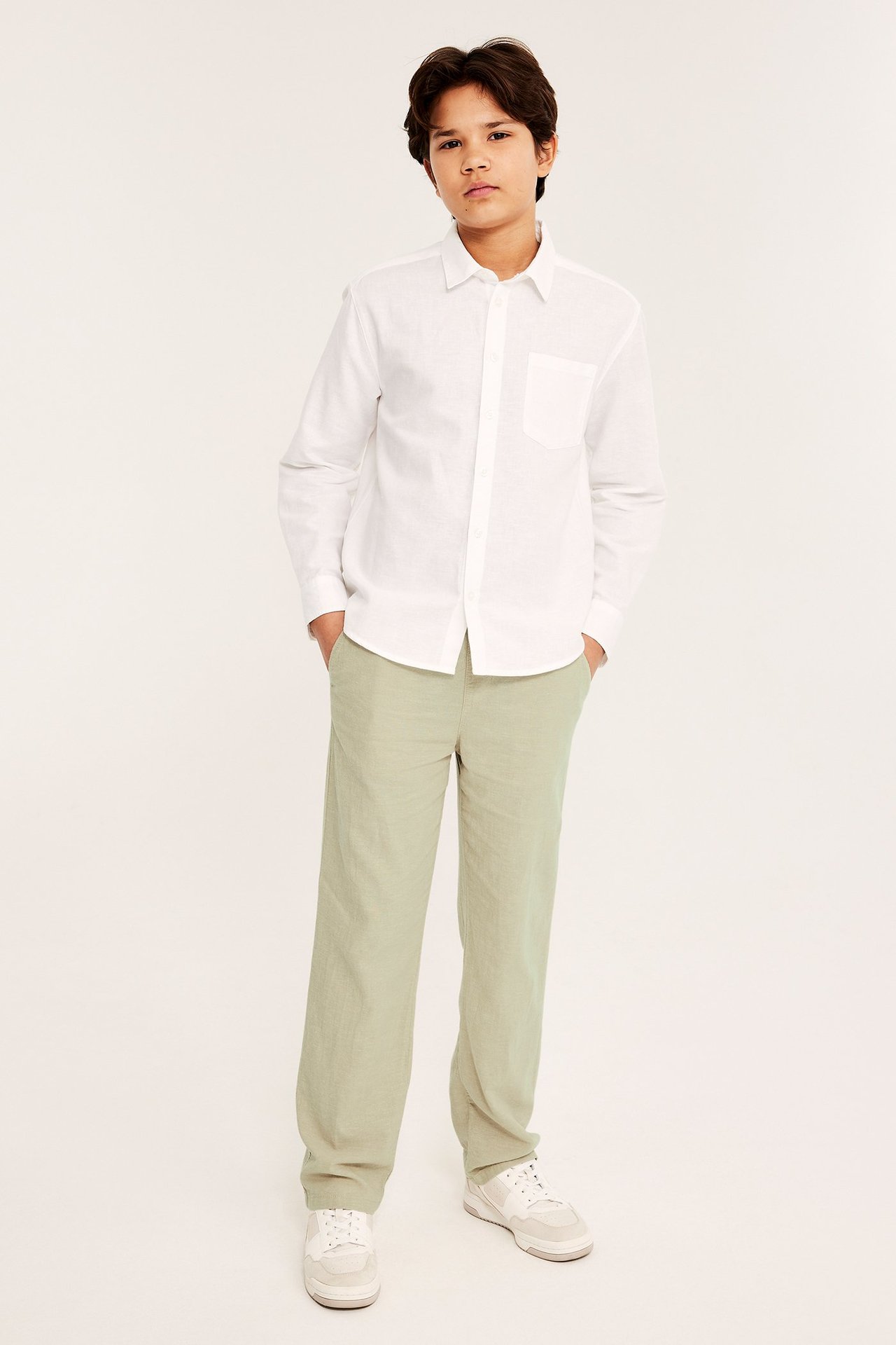 Edestä: lapsi väljissä vaaleanvihreissä pellavasekoitehousuissa, valkoisessa pellavapaidassa ja tennareissa.