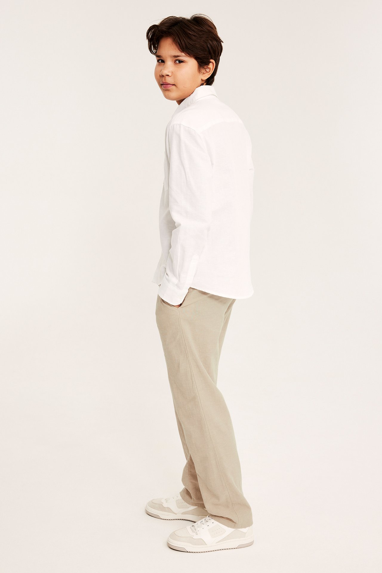 Widok z boku dziecka w beżowych spodniach z mieszanki lnu, białej koszuli i białych tenisówkach.