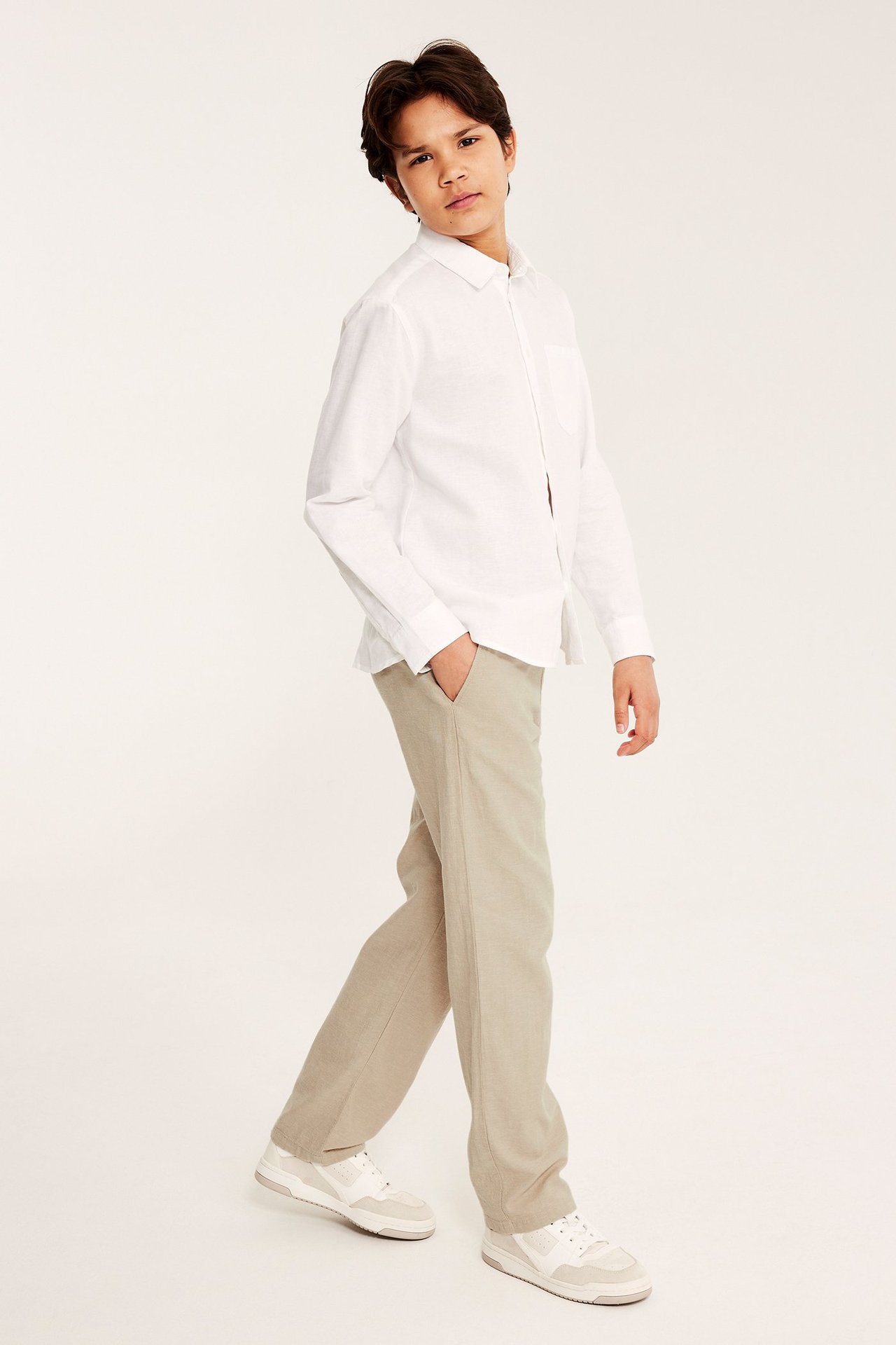 Framifrån, ett barn i lösa beiga linneblandade byxor, vit långärmad skjorta och vita sneakers.