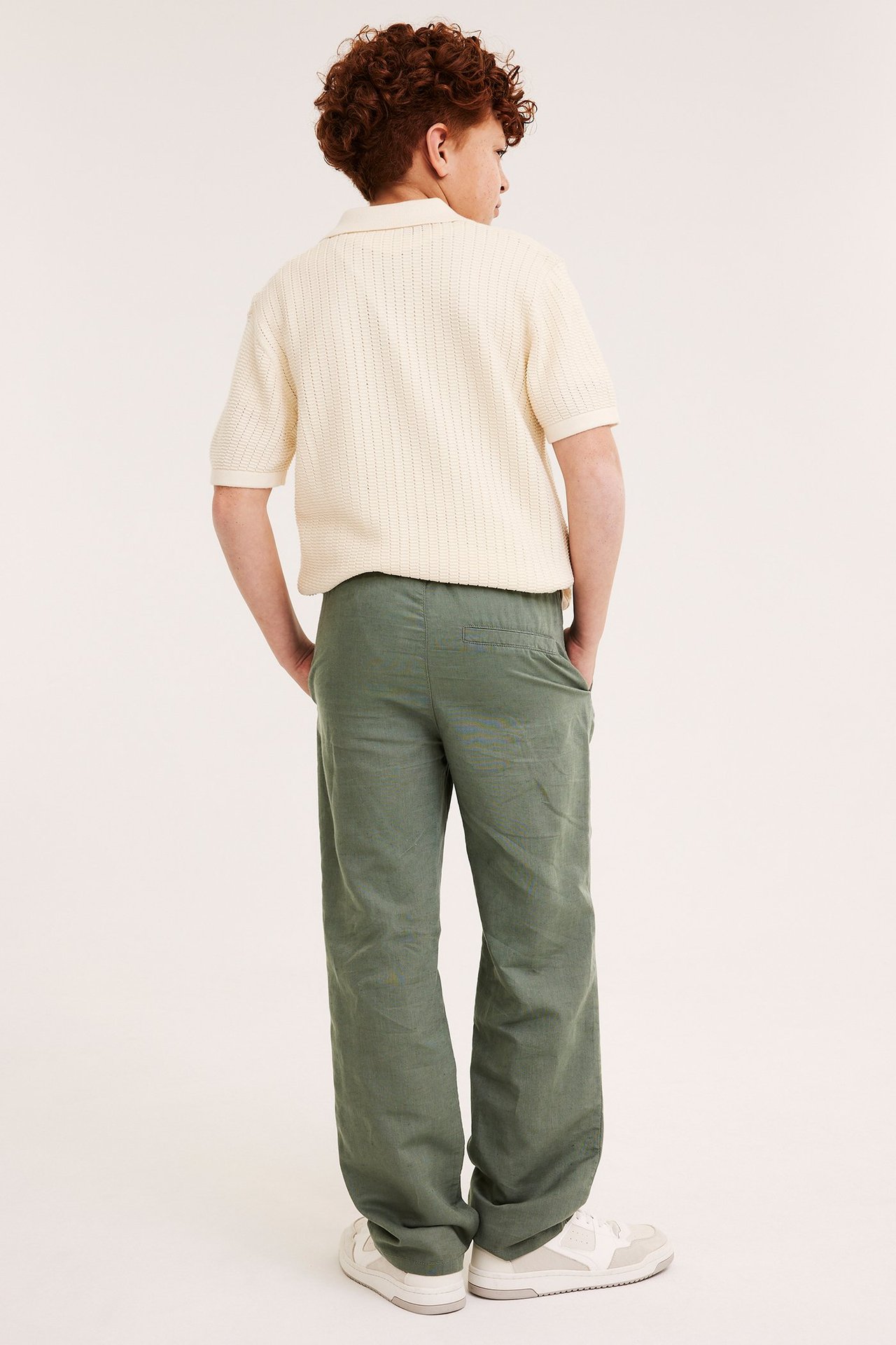 Widok z tyłu dziecka w zielonych luźnych lnianych spodniach, kremowej koszulce polo i białych tenisówkach.