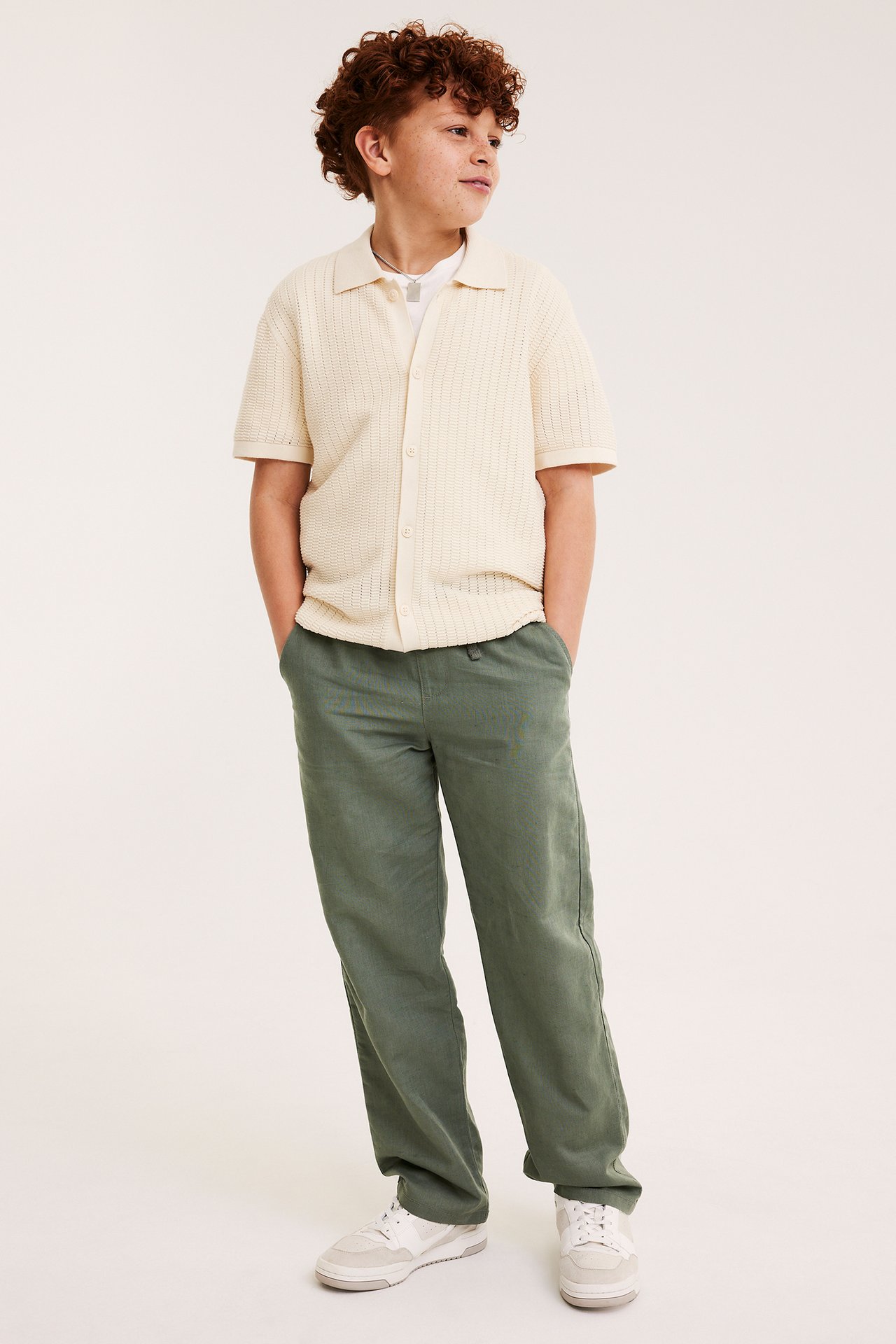 Framifrån, ett barn i gröna raka linneblandade byxor, en beige stickad pikétröja och vita sneakers.