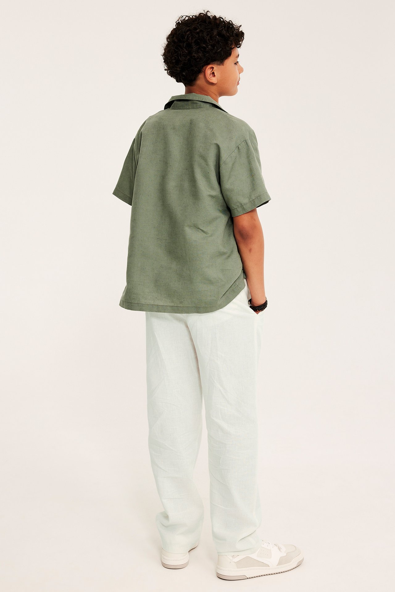Bakifrån: Kid bär lösa off-white linneblandade byxor med dragsko och en grön kortärmad skjorta.