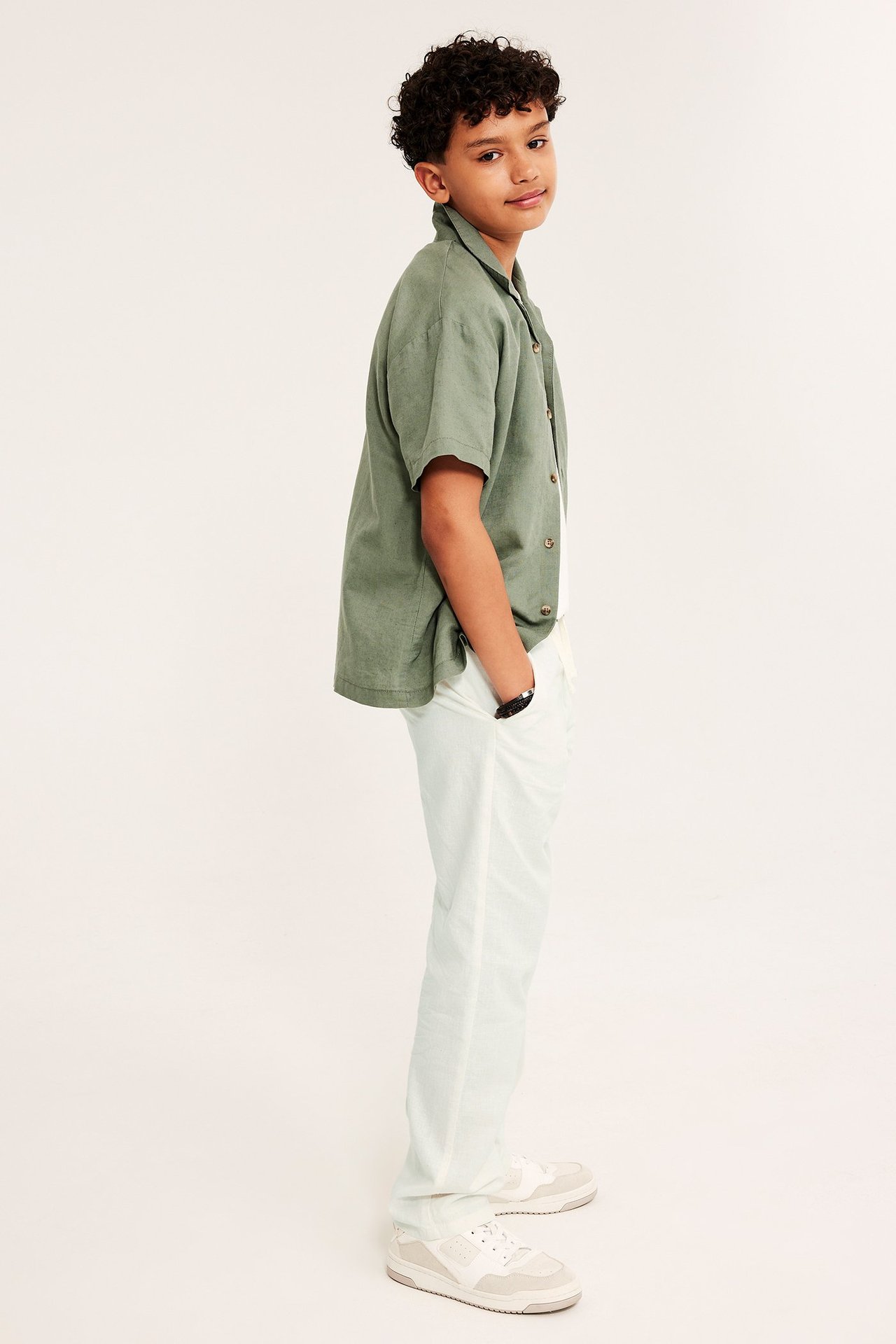 Sidovy av en pojke i avslappnade off-white byxor i linnemix, grön kortärmad skjorta och sneakers.