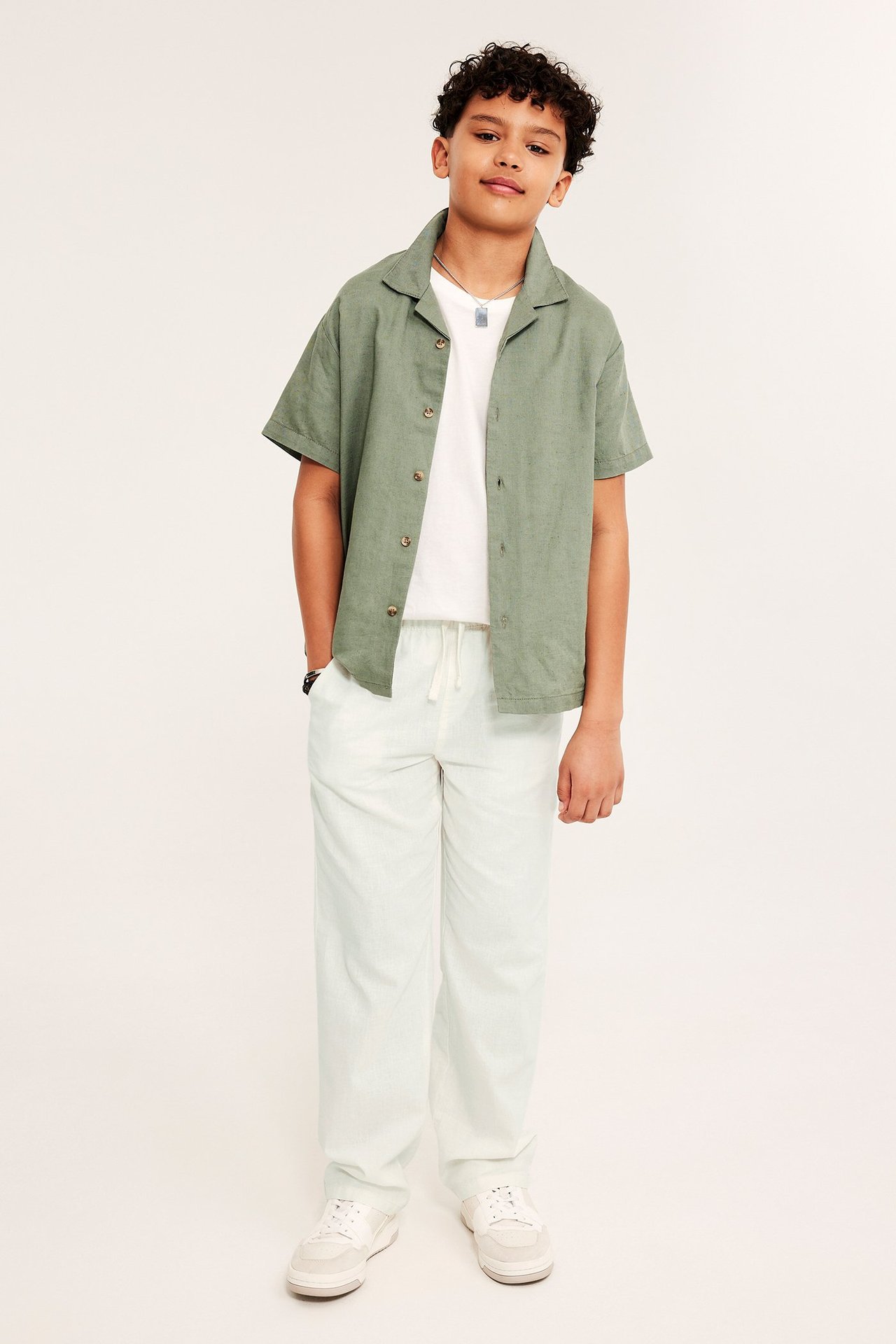 Framifrån, ett barn i offwhite linneblandade dragskobyxor, en grön skjorta och vita sneakers.