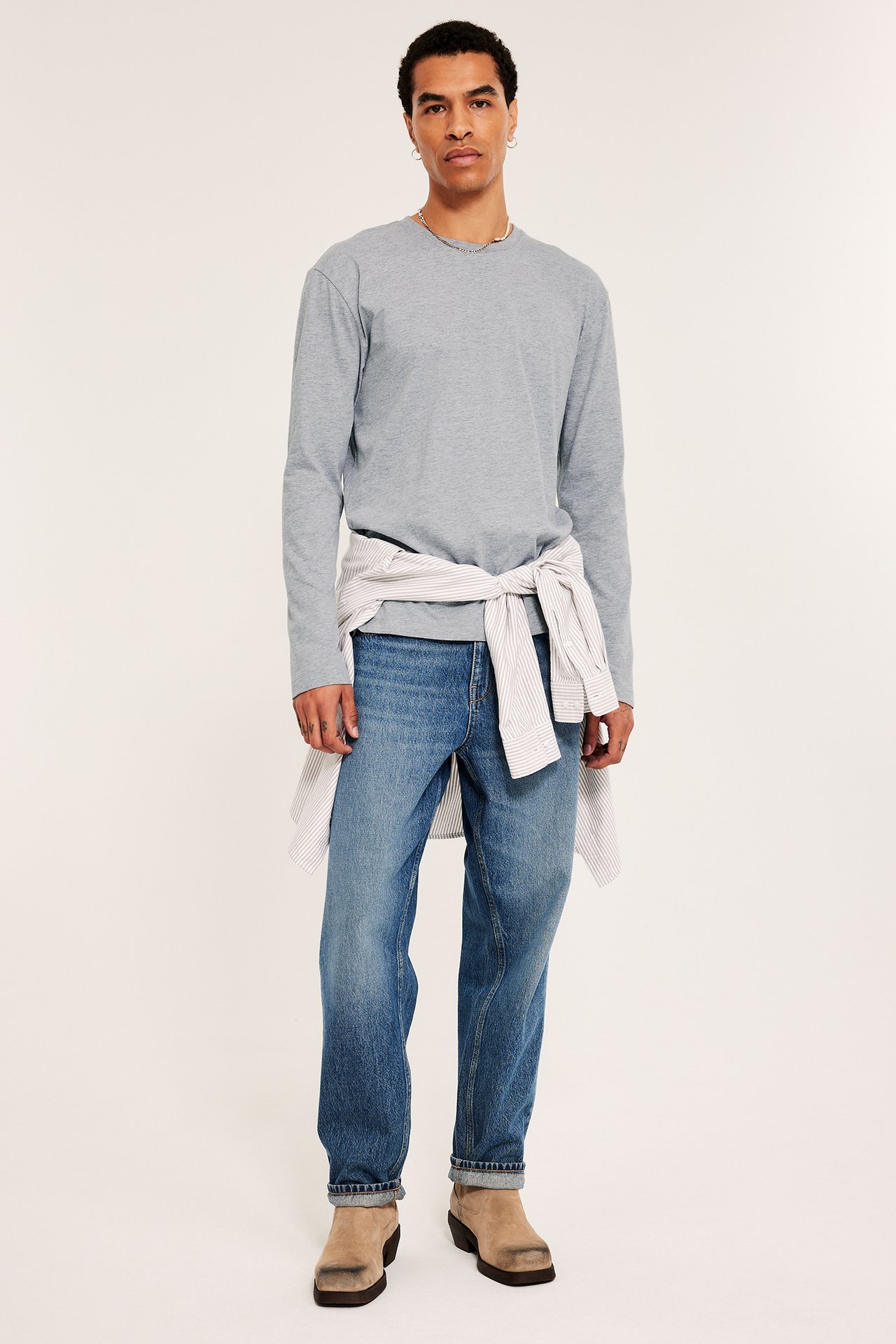 Mies yllään vaaleanharmaa pitkähihainen t-paita, siniset farkut ja raidallinen paita sidottuna vyötärölle.