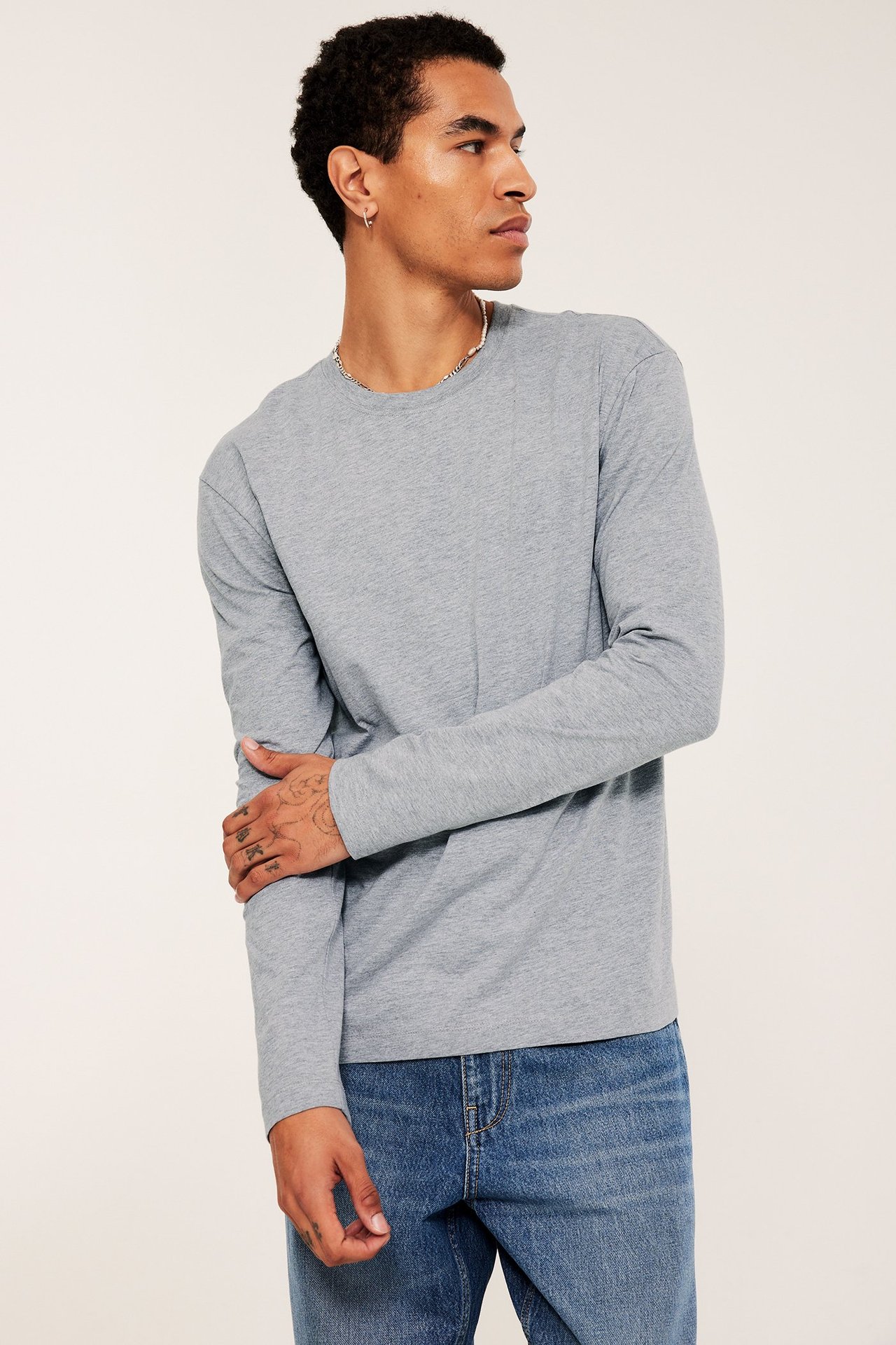 Mężczyzna w jasnoszarym longsleeve, niebieskich dżinsach, srebrnym naszyjniku i kolczyku, widok z przodu-boku.