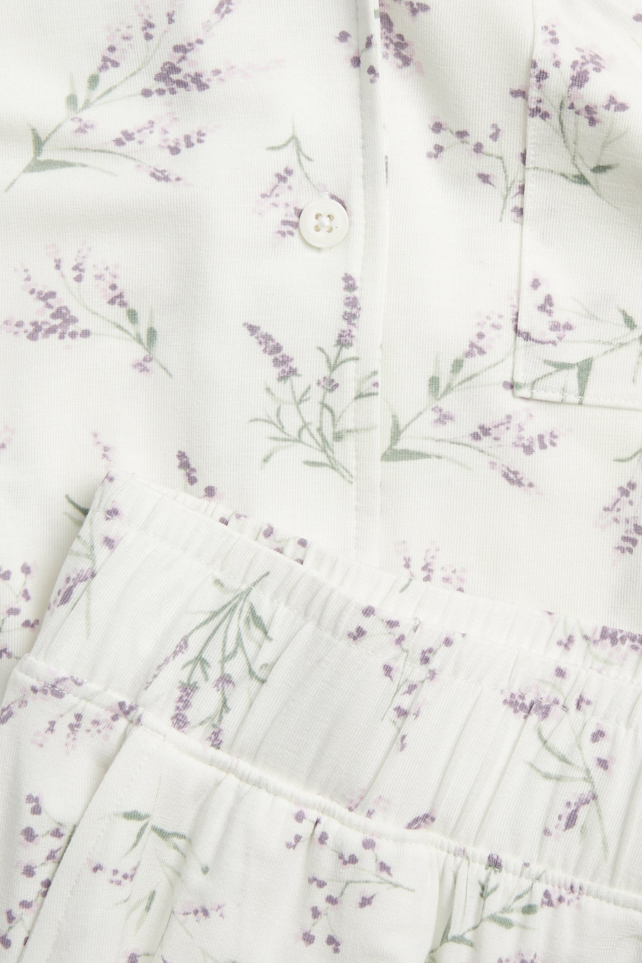 Nærbilde: hvitt lyocell-pyjamas med lys lilla blomstermønster. Skjorte med knapp og lomme, bukse med strikk.