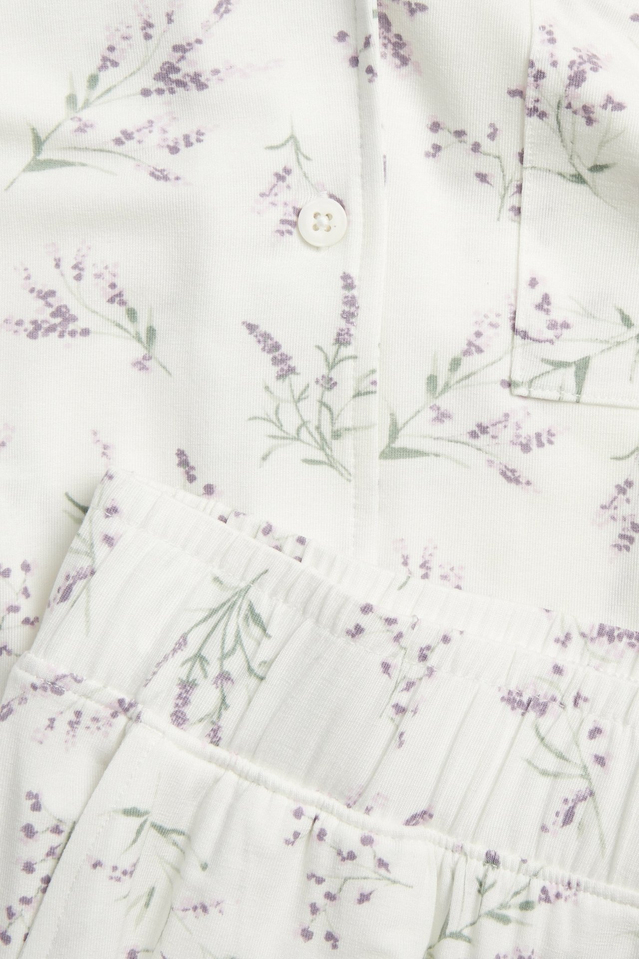Nærbilde: hvitt lyocell-pyjamas med lys lilla blomstermønster. Skjorte med knapp og lomme, bukse med strikk.
