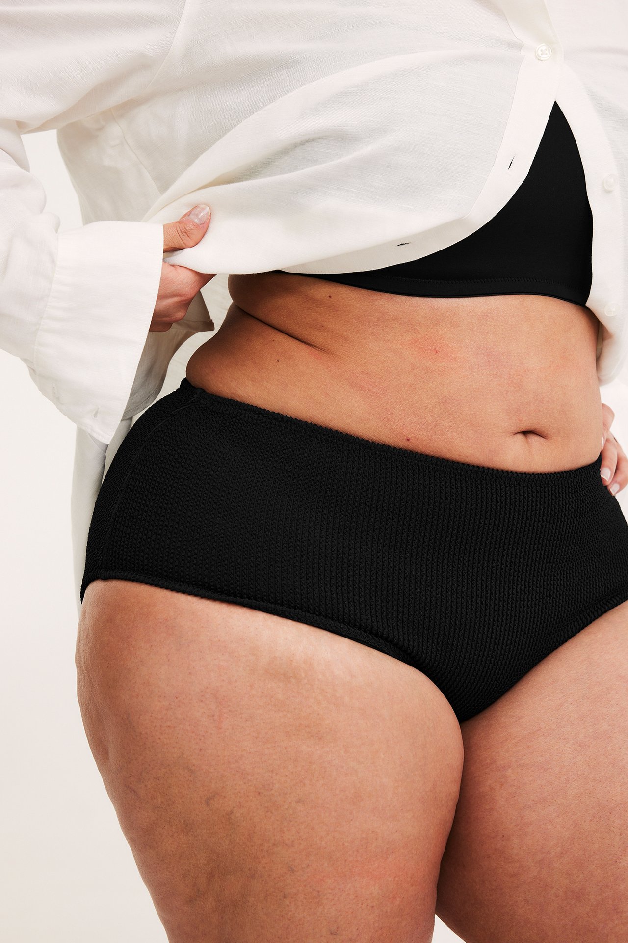 Sivukuva naisesta mustissa strukturoiduissa hipster-bikinihousuissa, musta toppi ja valkoinen paita.