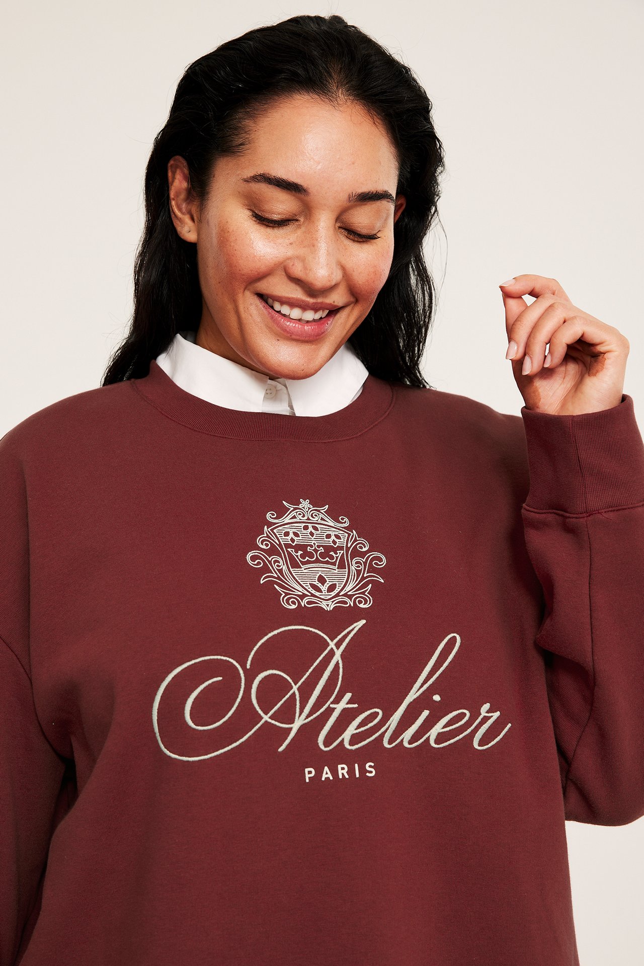 Nærbilde av kvinne i rød sweatshirt med hvitt emblem og 'Atelier Paris' broderi, over hvit skjorte.