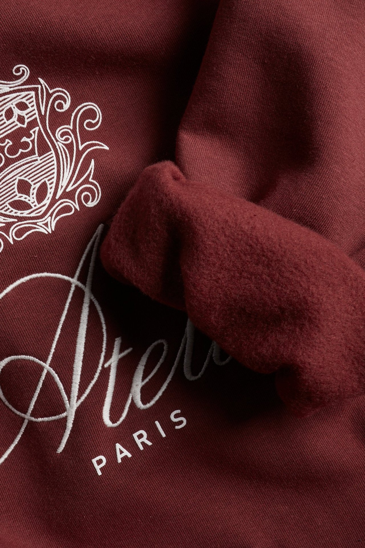 Zbliżenie czerwonej bluzy z białym haftem 'Atelier PARIS', nadrukiem emblematu i miękkim, szczotkowanym wnętrzem.