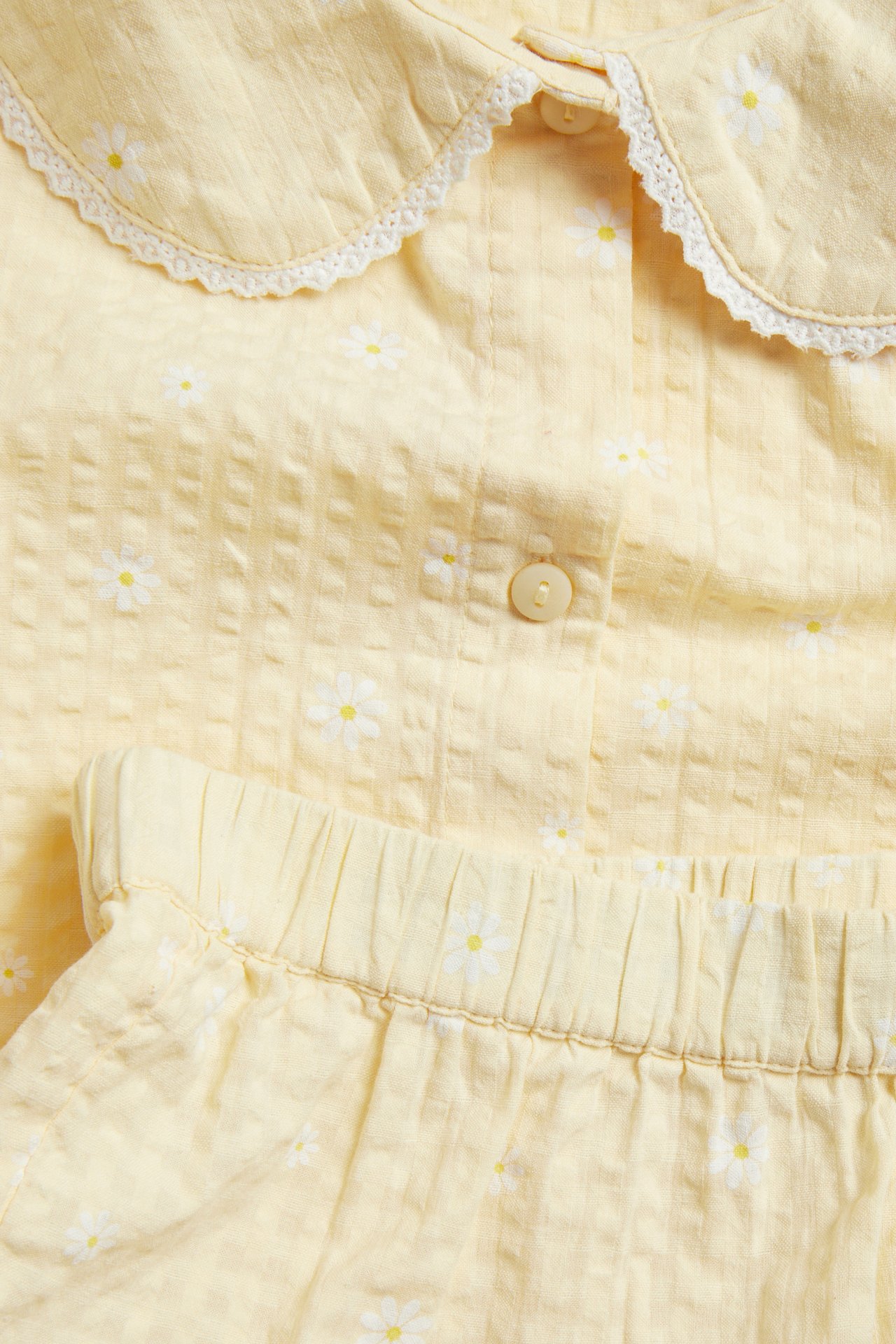Lähikuva vaaleankeltaisesta päivänkakkara-kuvioidusta bäckebölja-pyjamasettistä, jossa pitsikaulus ja joustava vyötärö.
