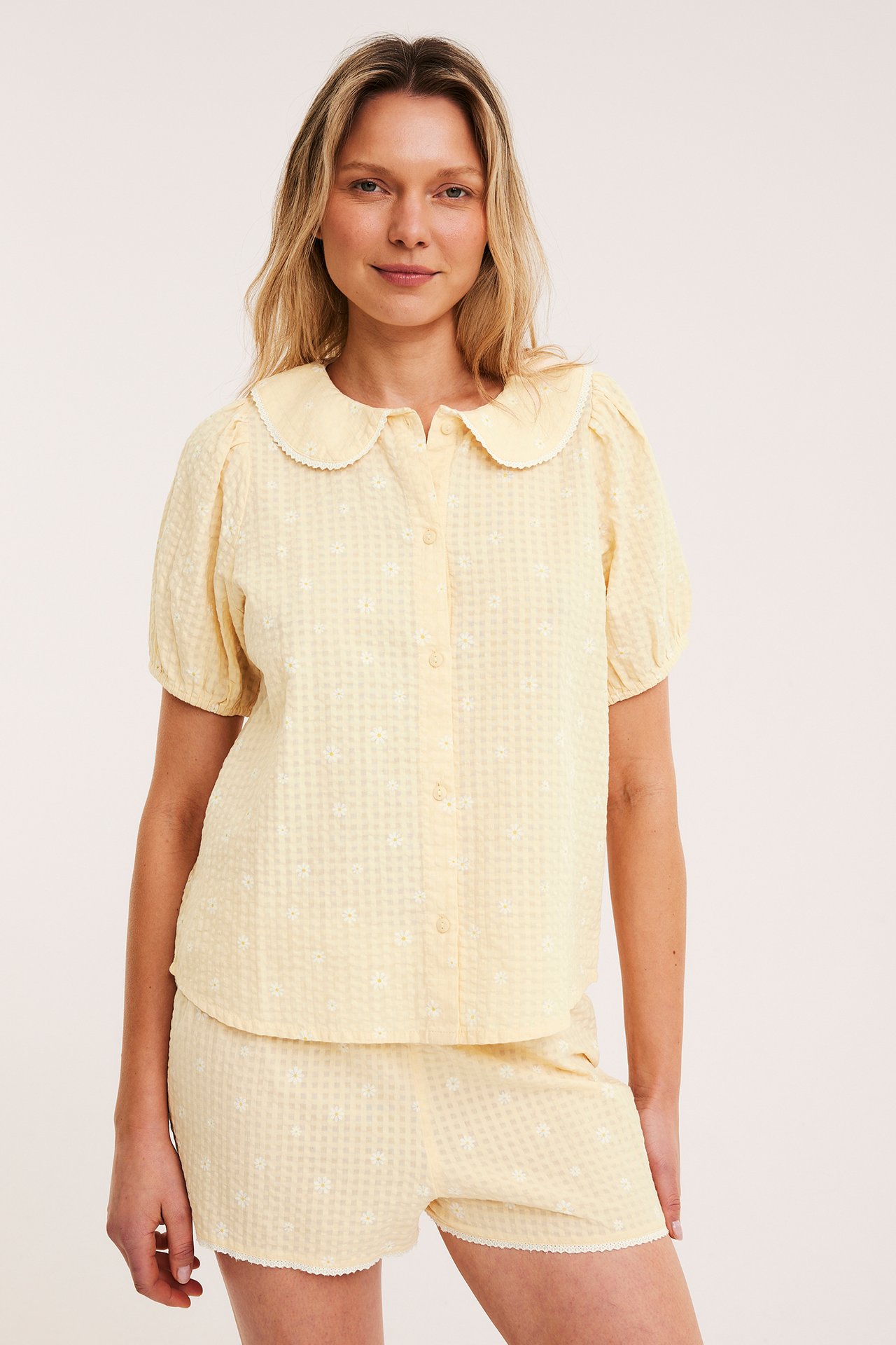 Frontvy av kvinna i ljusgult blommigt bäckeböljapyjamas: topp med spetskrage och puffärm, matchande shorts.