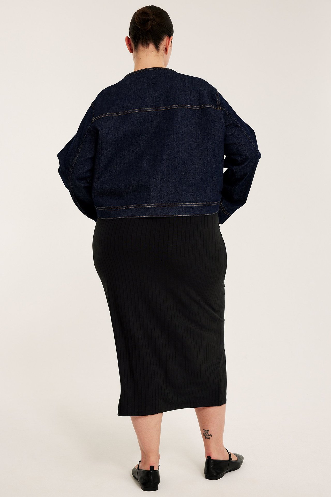 Bakifrån en kvinna i en svart lång ribbad jerseykjol med resårmidja och en mörkblå jeansjacka.