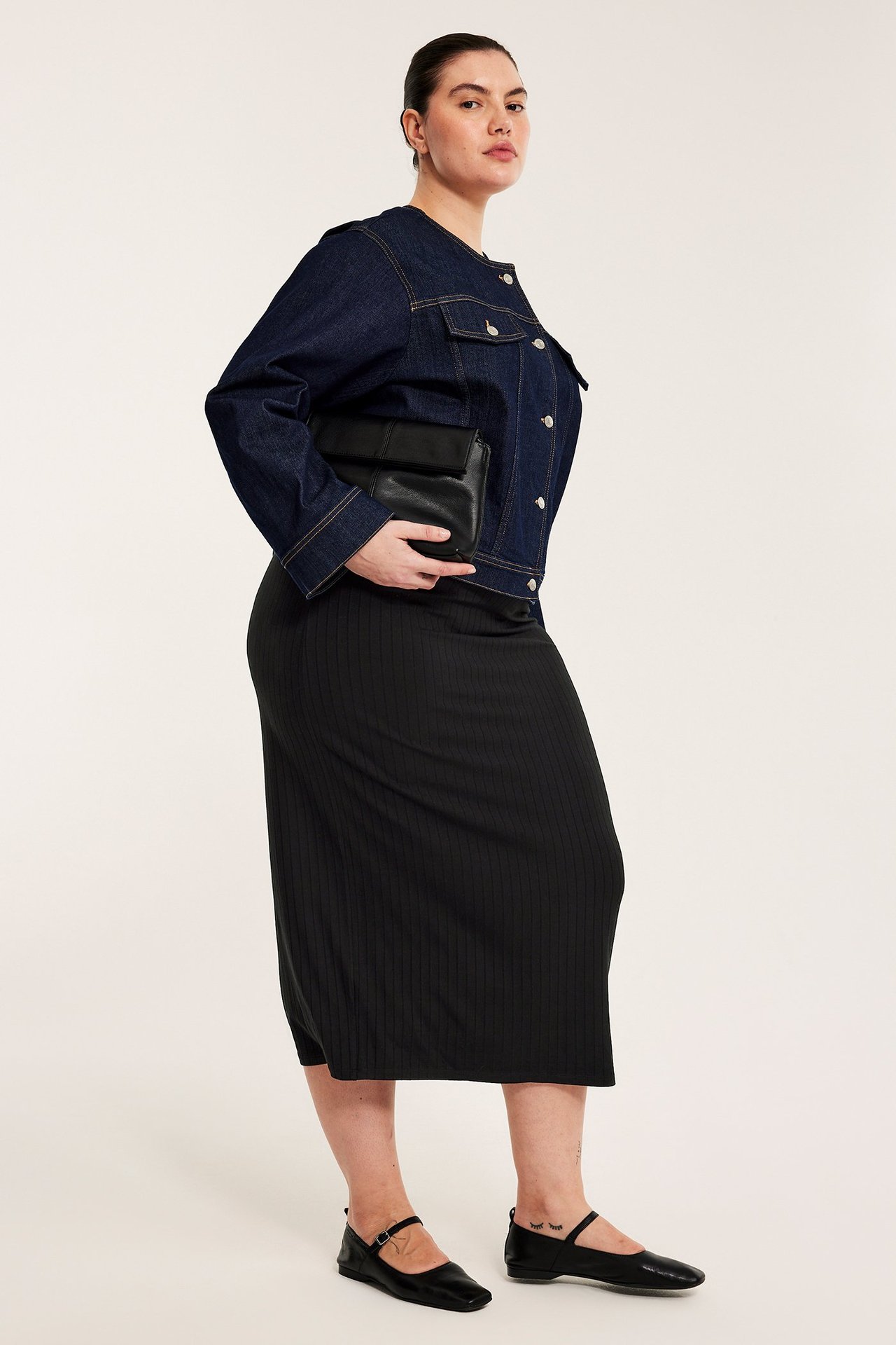 Sidovy av kvinna i lång svart ribbad kjol, mörkblå jeansjacka, med svart clutch och svarta ballerinaskor.