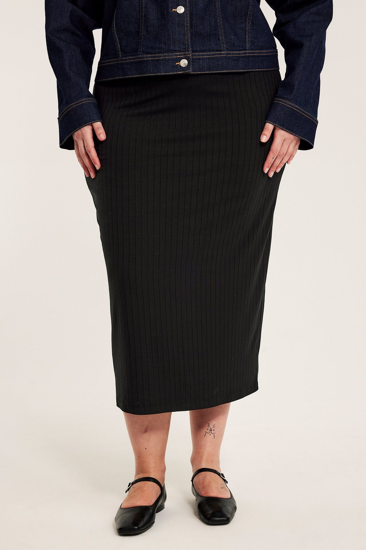 Framifrån: kvinna i lång svart ribbad jerseykjol, mörkblå jeansjacka och svarta platta skor.