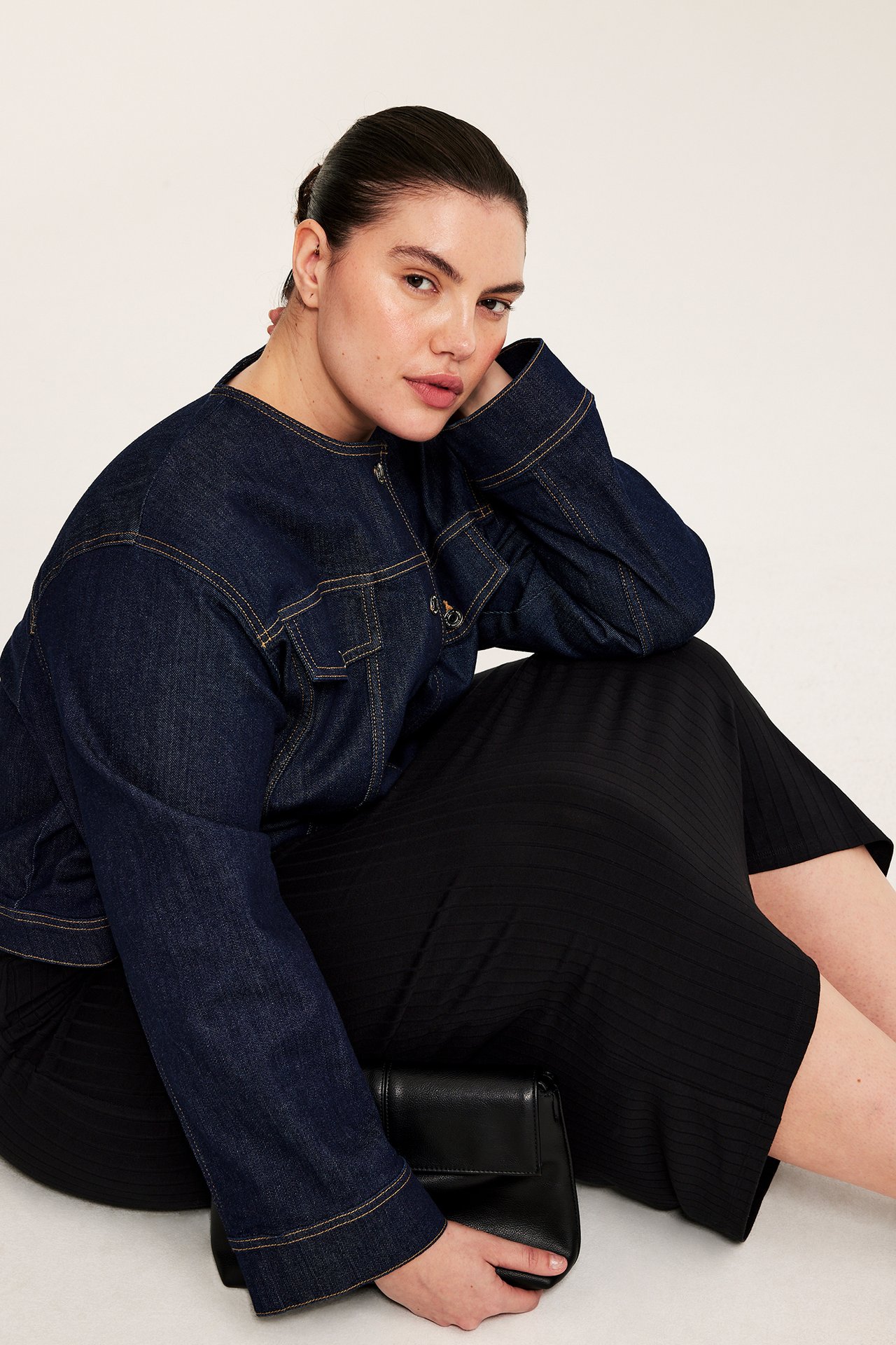 Framifrån: kvinna i svart lång ribbad jerseykjol, mörkblå jeansjacka, håller en svart clutchväska.