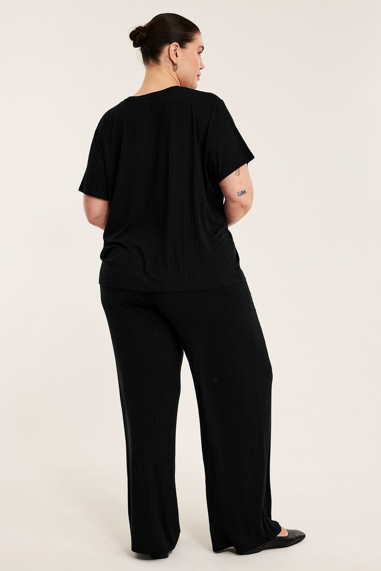 Widok z tyłu: kobieta w czarnych prążkowanych szerokich spodniach z wiskozy, topie i baletkach.