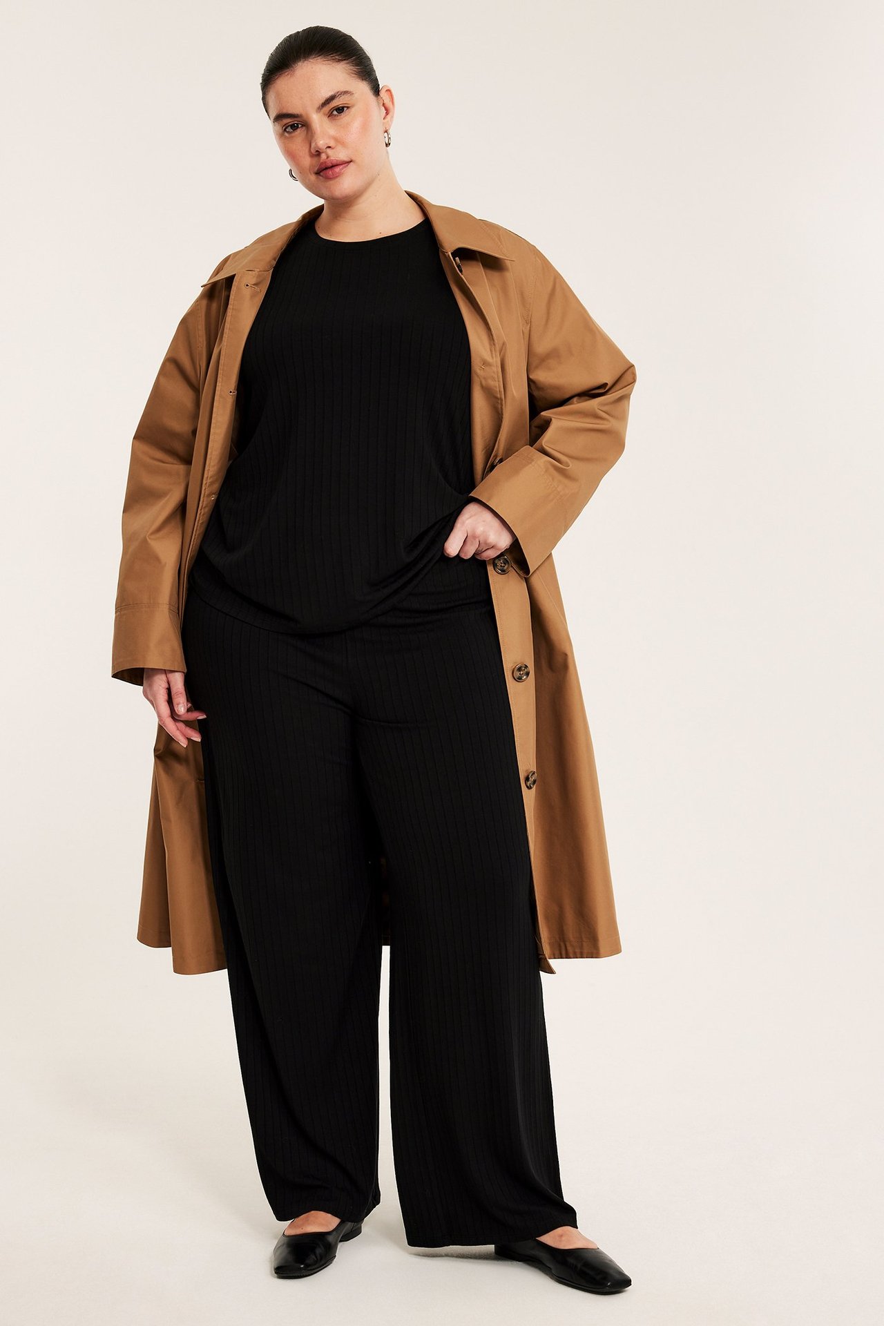 Frontbilde av en kvinne i svarte ribbestrikkede vide bukser, svart topp og brun trenchcoat.