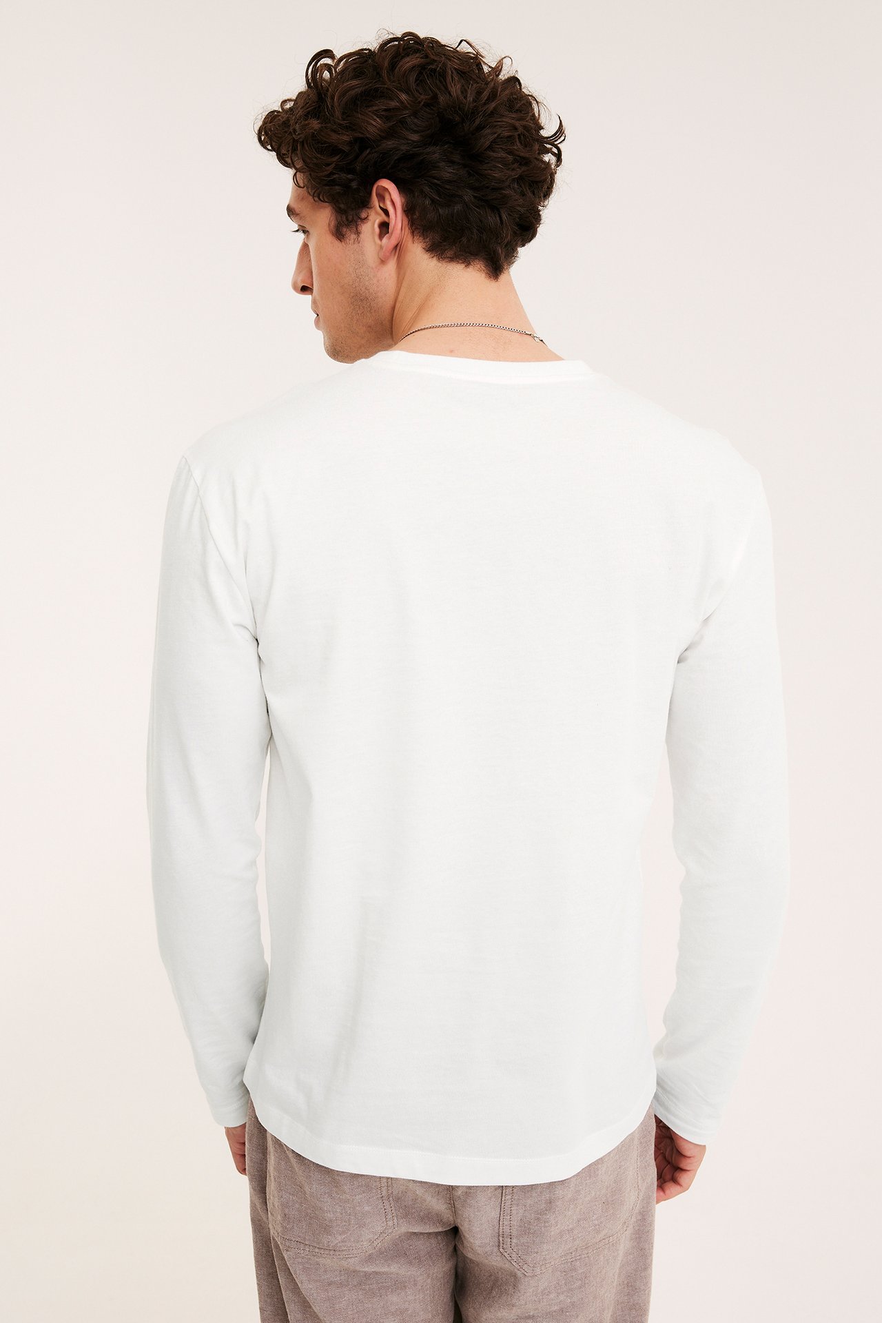 Widok z tyłu: mężczyzna w gładkim, ecru bawełnianym t-shircie z długim rękawem, brązowych spodniach i srebrnym łańcuszku.