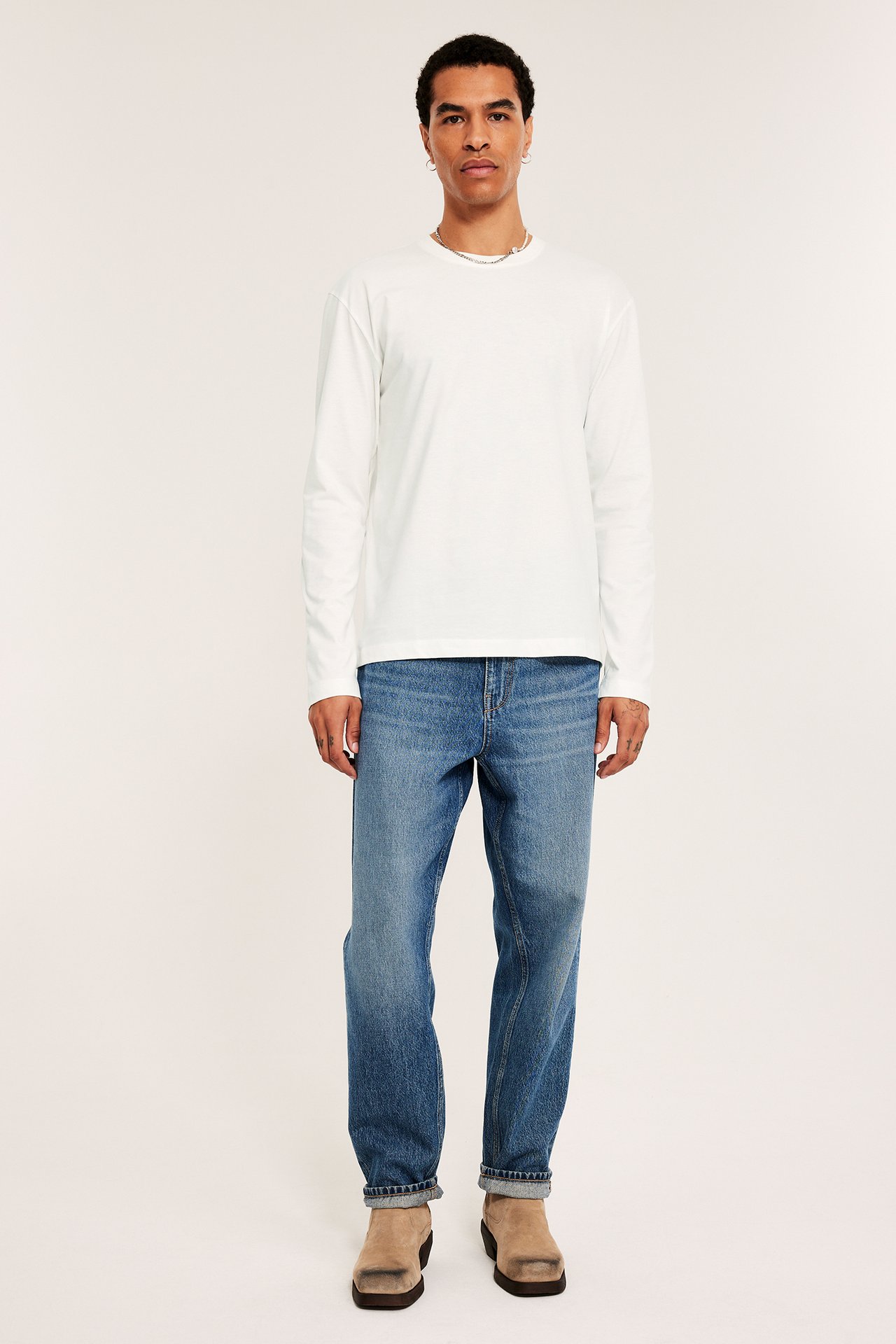 Mies edestäpäin yllään luonnonvalkoinen pitkähihainen T-paita, siniset farkut ja beiget nilkkurit.