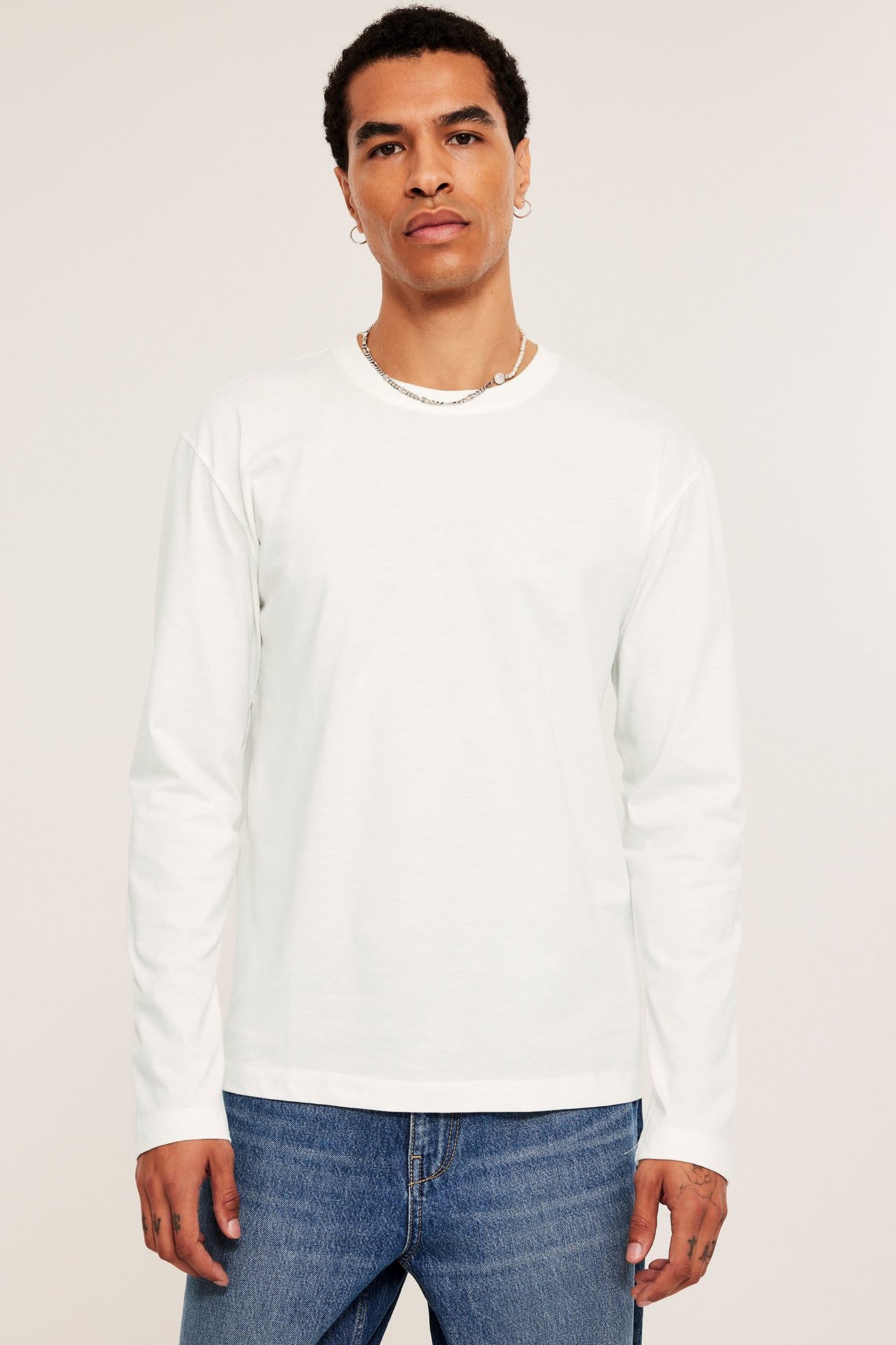 Mężczyzna od przodu w gładkiej, ecru, bawełnianej koszulce z długim rękawem i niebieskich dżinsach.