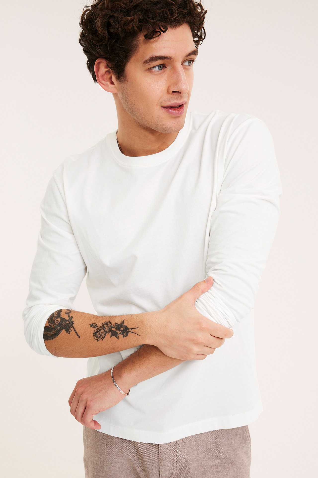 Widok z przodu: mężczyzna w bawełnianej koszulce z długim rękawem ecru, jasnobrązowych spodniach i srebrnej bransoletce.