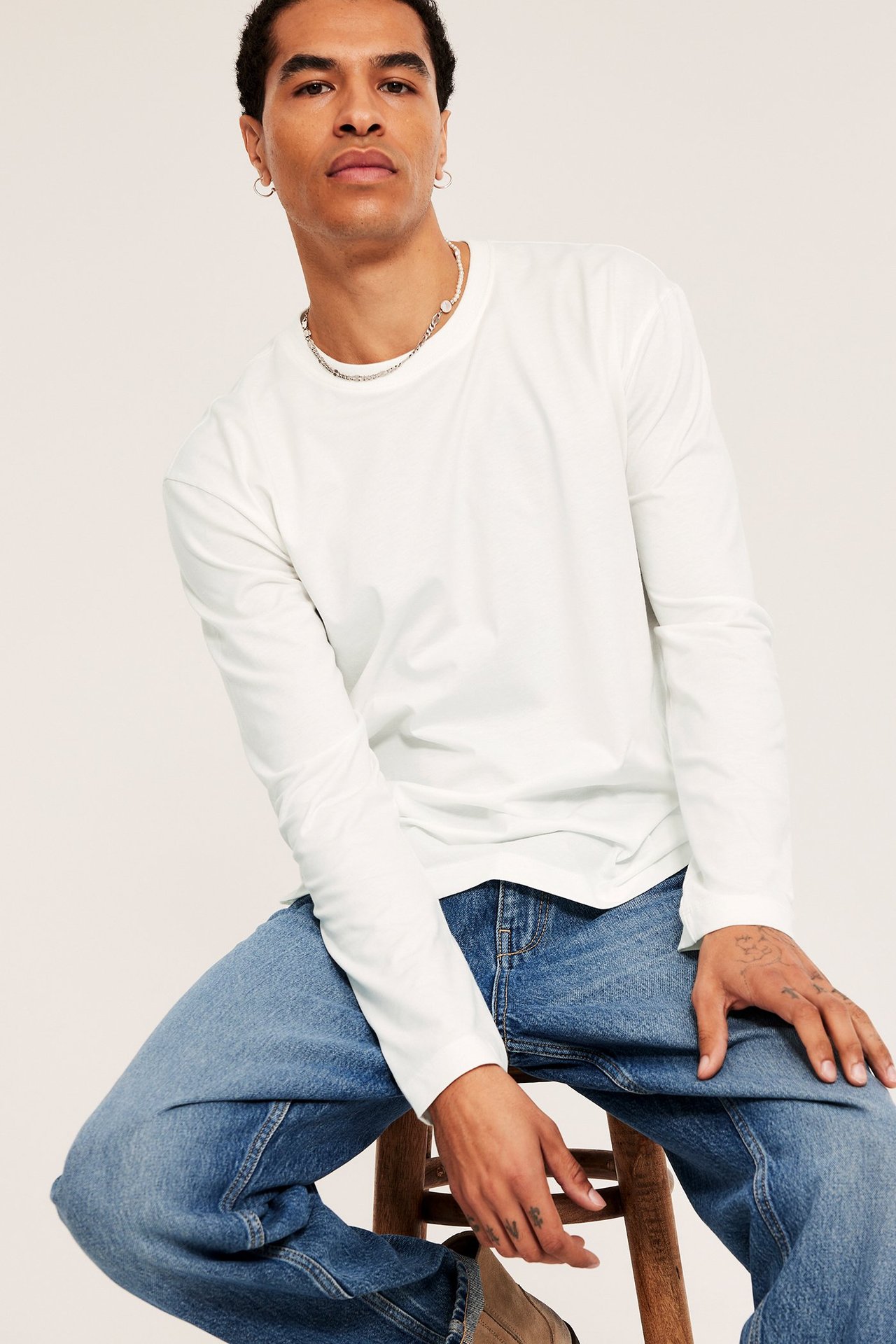 Widok z przodu mężczyzny w gładkiej koszulce z długim rękawem ecru, niebieskich dżinsach i srebrnym naszyjniku.
