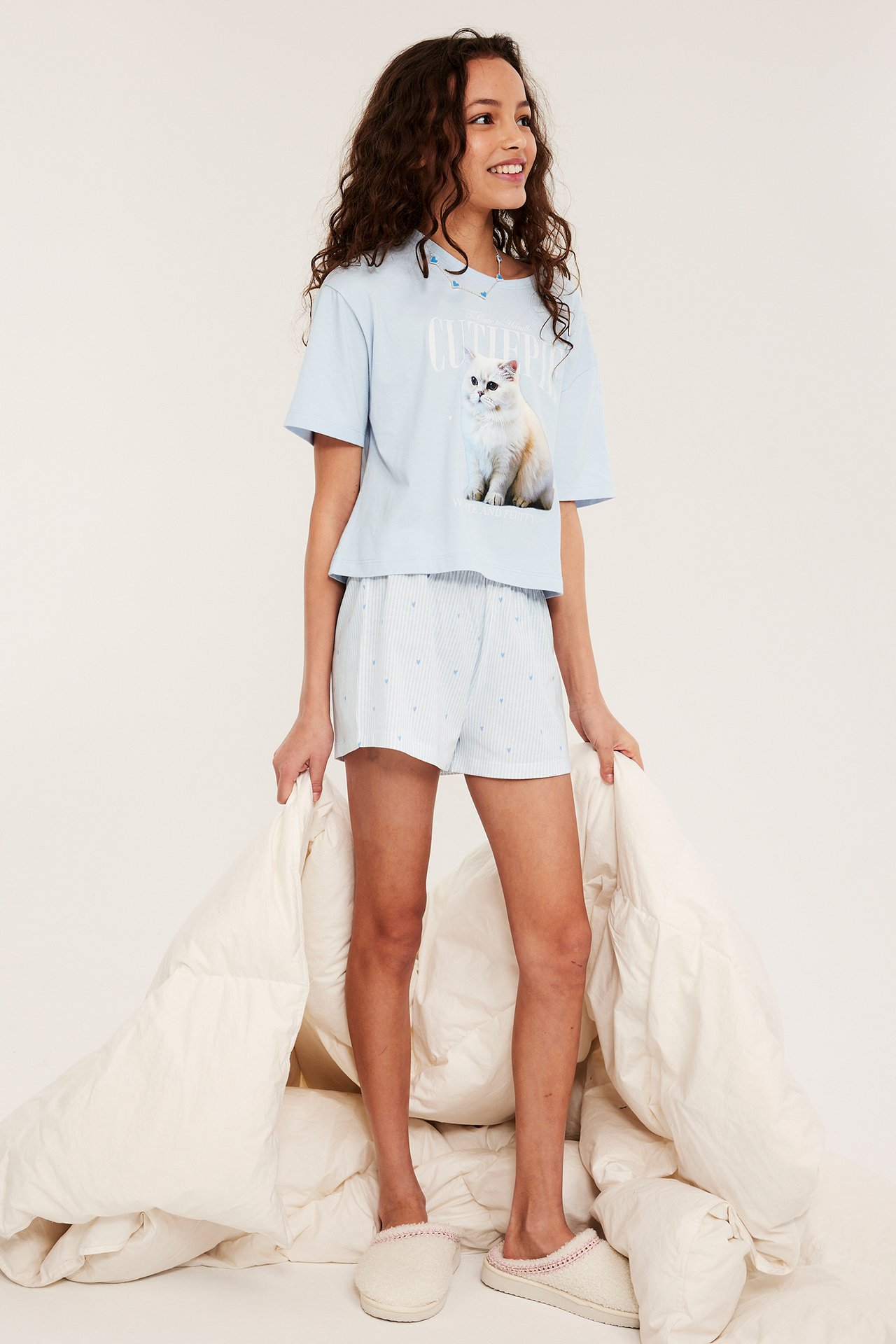 Forfra: Et barn i lysblå bomullspyjamas med kattetrykt t-skjorte, stripete shorts, beige tøfler, holder en dyne.