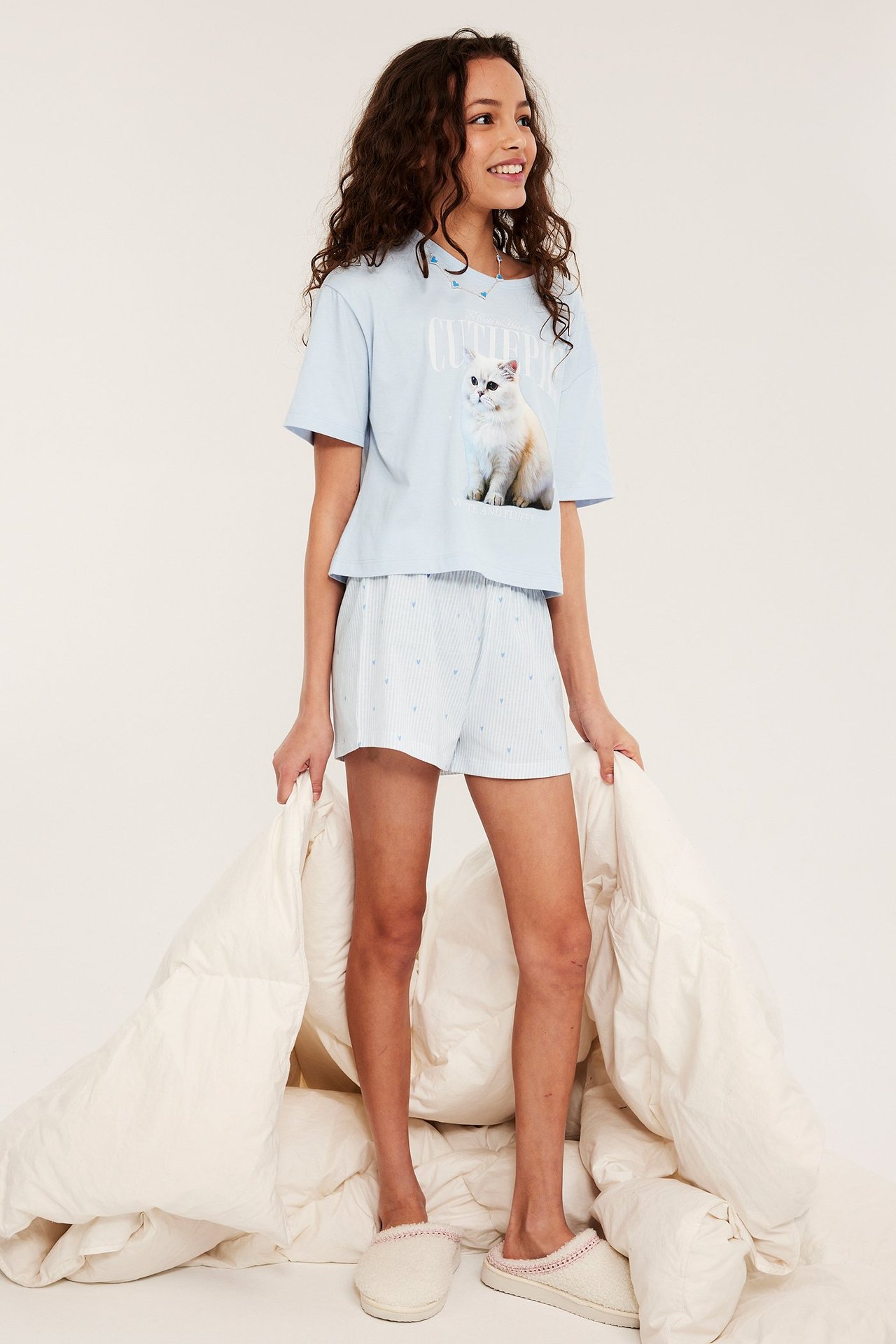 Edestä: Lapsi vaaleansinisessä kissapainatuspuuvillapyjamassa, raidalliset shortsit, beiget tossut, pitää peittoa.