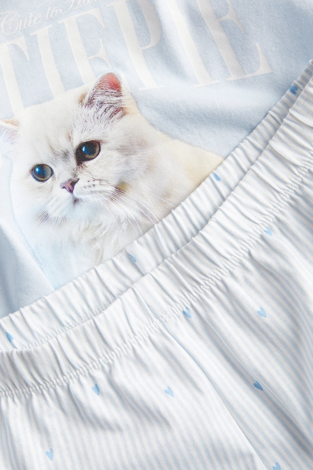 Zbliżenie: jasnoniebieska góra piżamy z nadrukiem białego kota i paski szorty z małymi niebieskimi sercami.