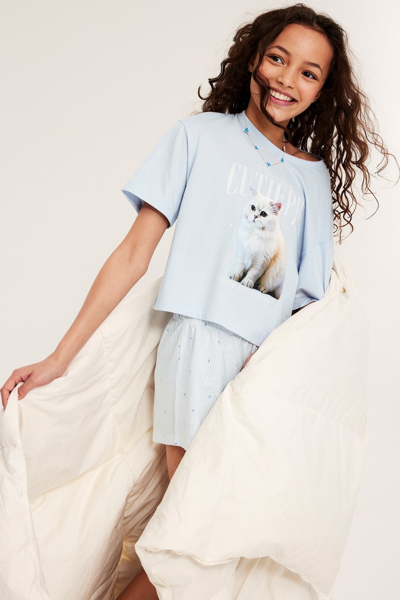 Widok z przodu: uśmiechnięte dziecko w jasnoniebieskiej piżamie z kotem, krótkie spodenki, trzyma kołdrę.