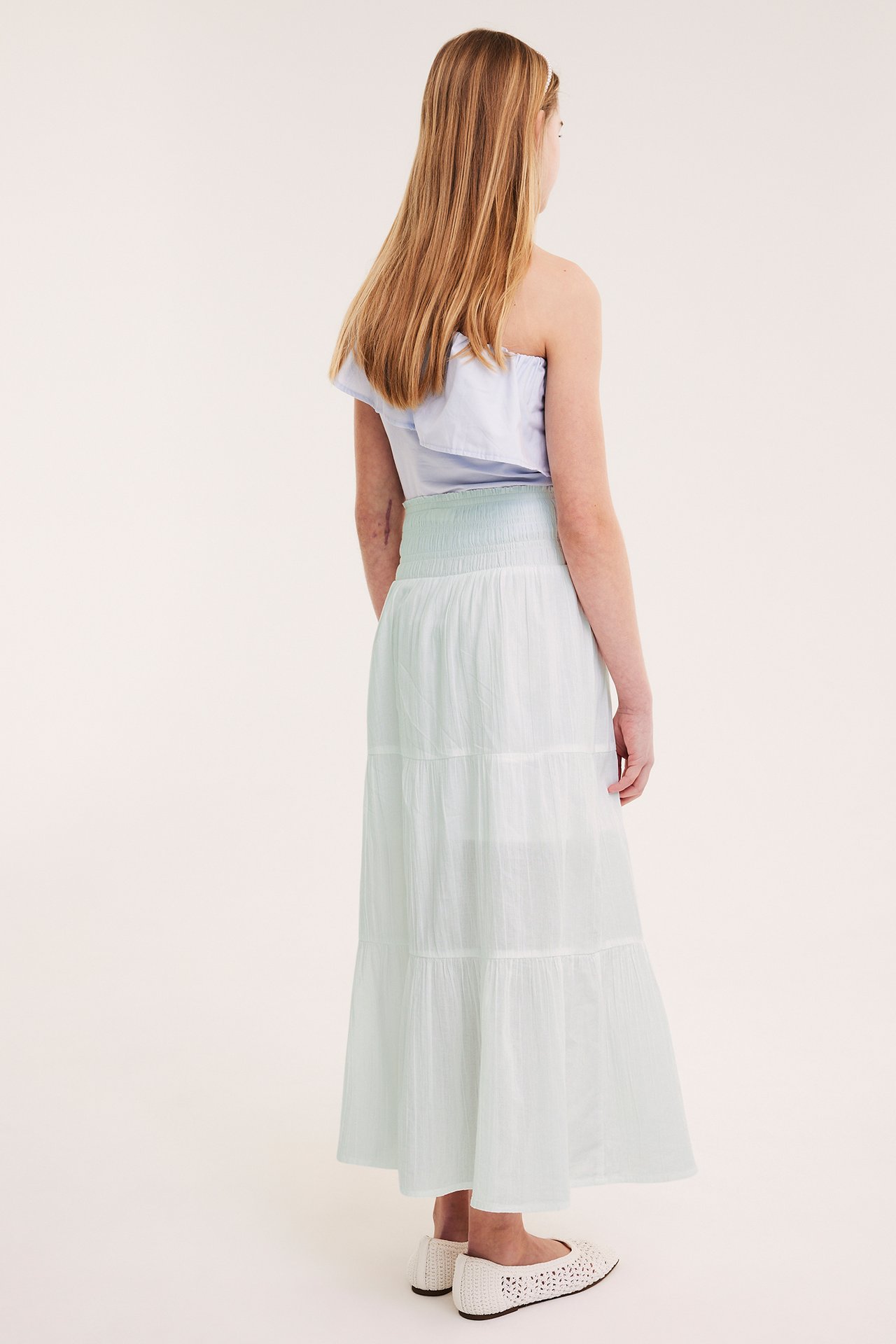 Dziecko, tyłem: jasnozielona długa spódnica z marszczoną talią, jasnoniebieski top, białe buty.
