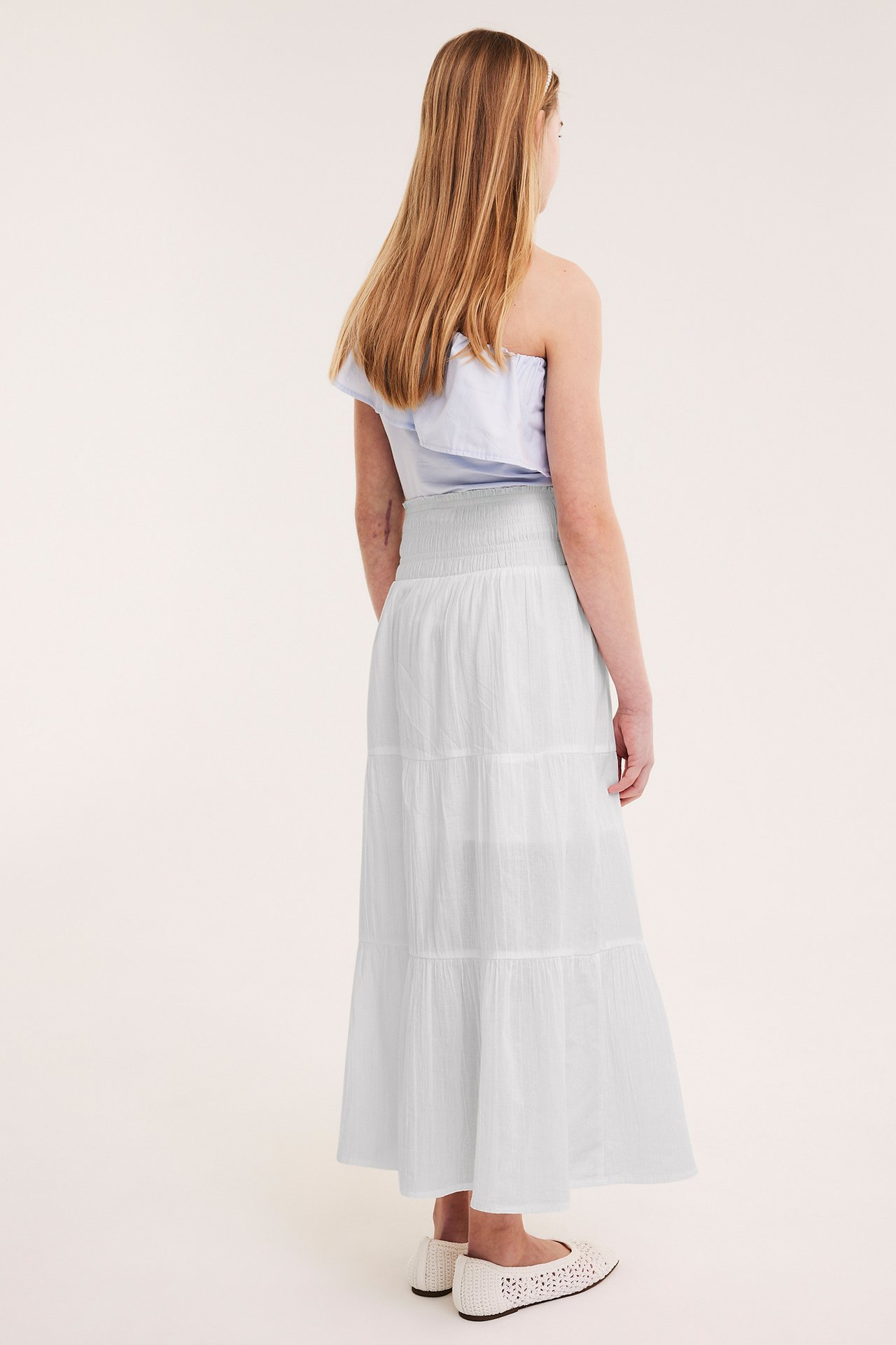 Widok z tyłu dziecka ubranego w białą bawełnianą spódnicę maxi, jasnoniebieski top na jedno ramię i białe baleriny.
